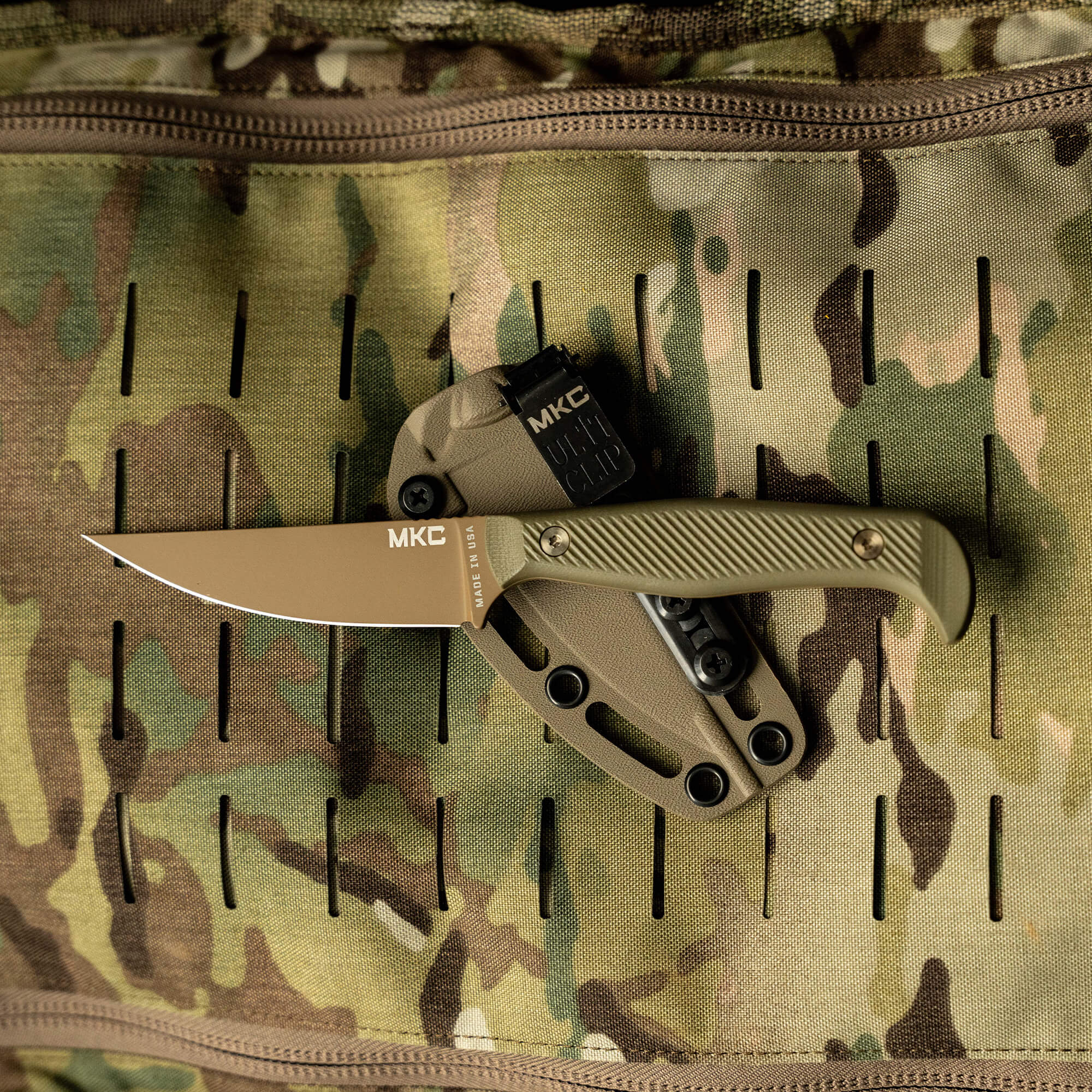 Alt text: "Close-up of a tan and olive green folding knife marked 'MKC' on a camouflage background with a tactical Molle system, highlighting the blade and textured handle."