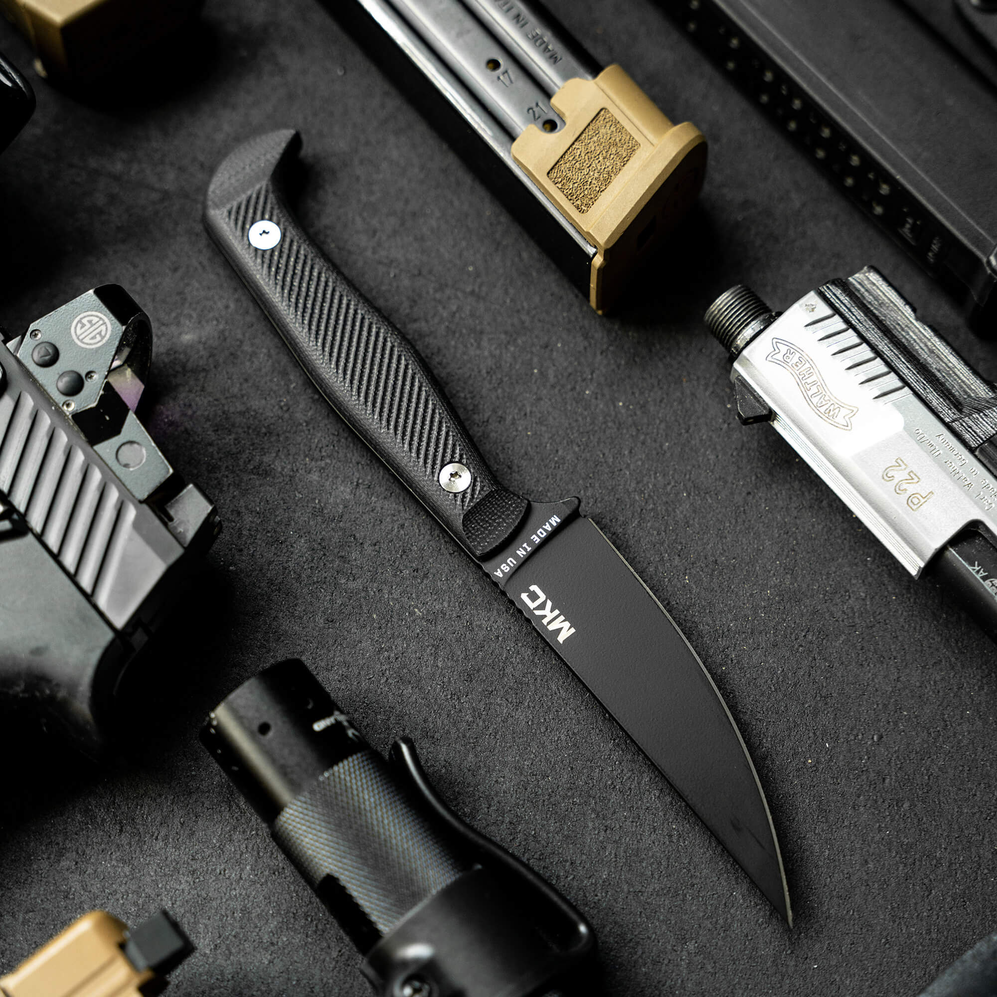 Alt text: High-quality black MKM battle knife with a textured handle displayed alongside various tactical gear including ammunition clips, a brass knuckle, and a multi-tool, all arranged on a dark textured surface.