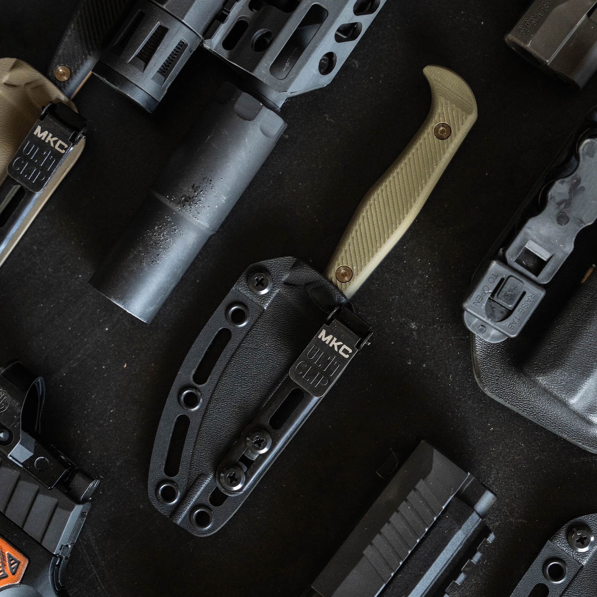 Alt text: "Close-up of a tactical knife, model 'BATTLE GOAT - BLK/OD', secured in a black sheath surrounded by various tactical gear and firearm accessories on a dark surface."
