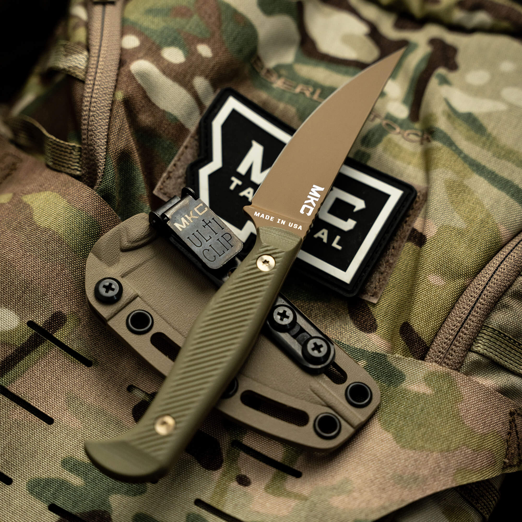 Alt text: "Close-up image of the BATTLE GOAT folding knife in coyote olive drab color. The knife is partially open, showing a sharp, tan blade and a textured handle, resting on a camouflaged tactical gear fabric with