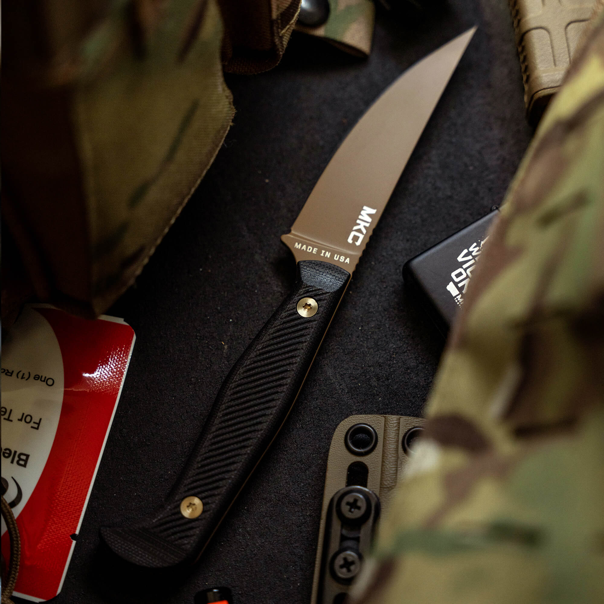 Alt text: "Close-up of a BATTLE GOAT tactical knife in coyote black with a textured handle, prominently displaying the brand and 'Made in USA' on the blade, positioned among tactical gear and camouflage materials."