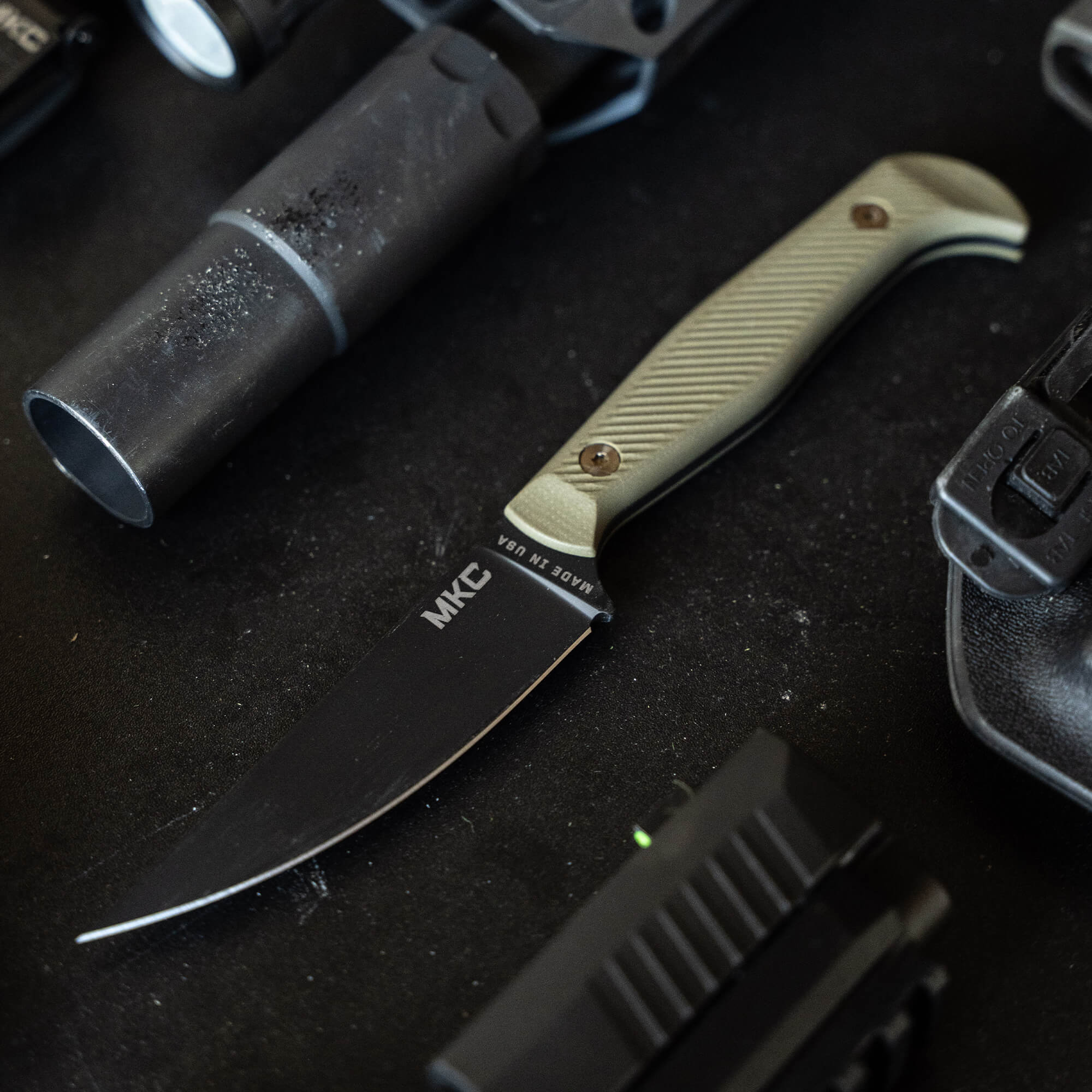 Alt text: "Close-up image of a tactical knife labeled MKC with a black blade and olive drab handle, surrounded by assorted military gear including a silencer and flashlight on a dark surface."