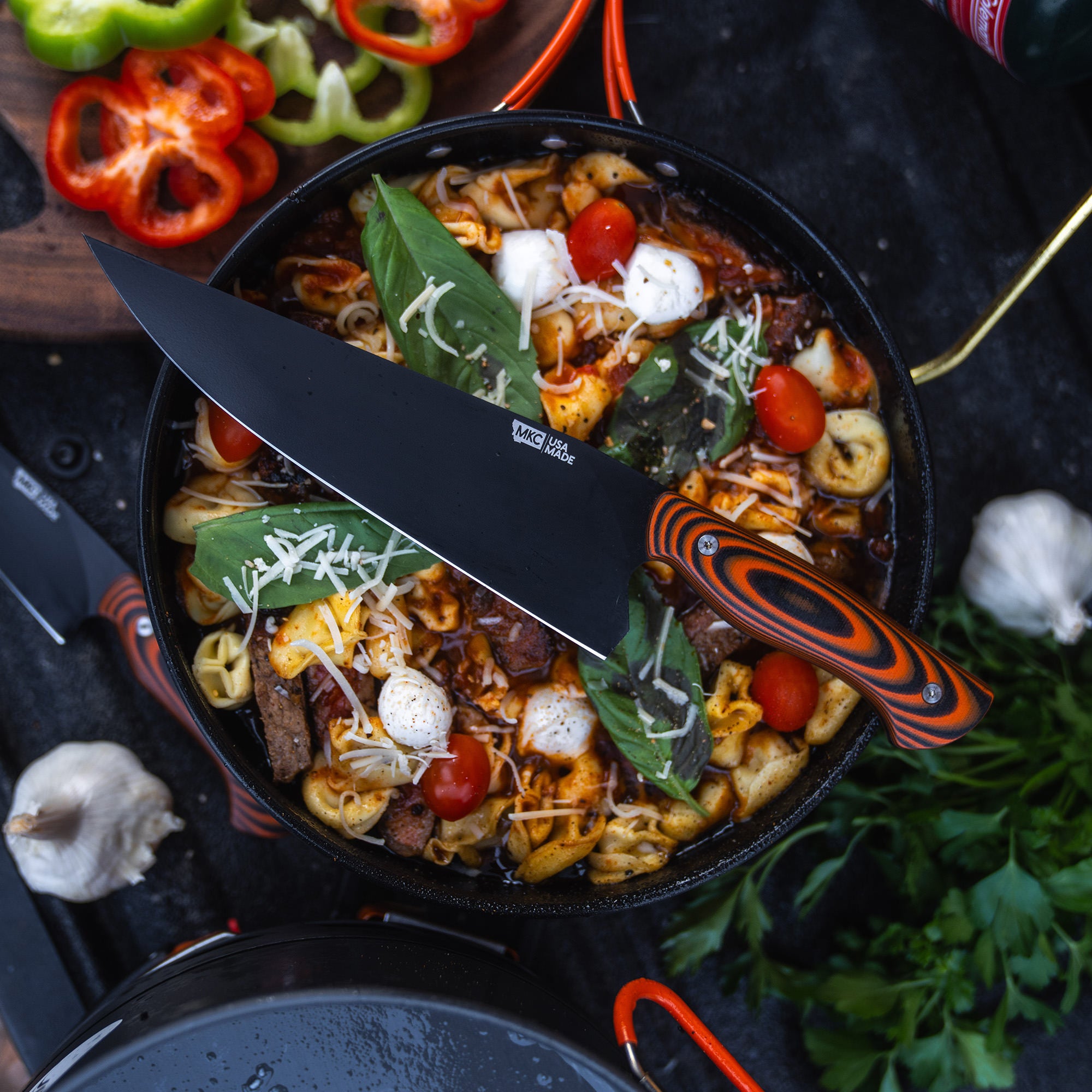 Alt text: "A vibrant cooking scene with a BIGHORN CHEF orange and black handled chef's knife resting on a skillet containing a colorful pasta dish with cherry tomatoes, basil and shredded cheese, surrounded by fresh ingredients including bell peppers, garlic,