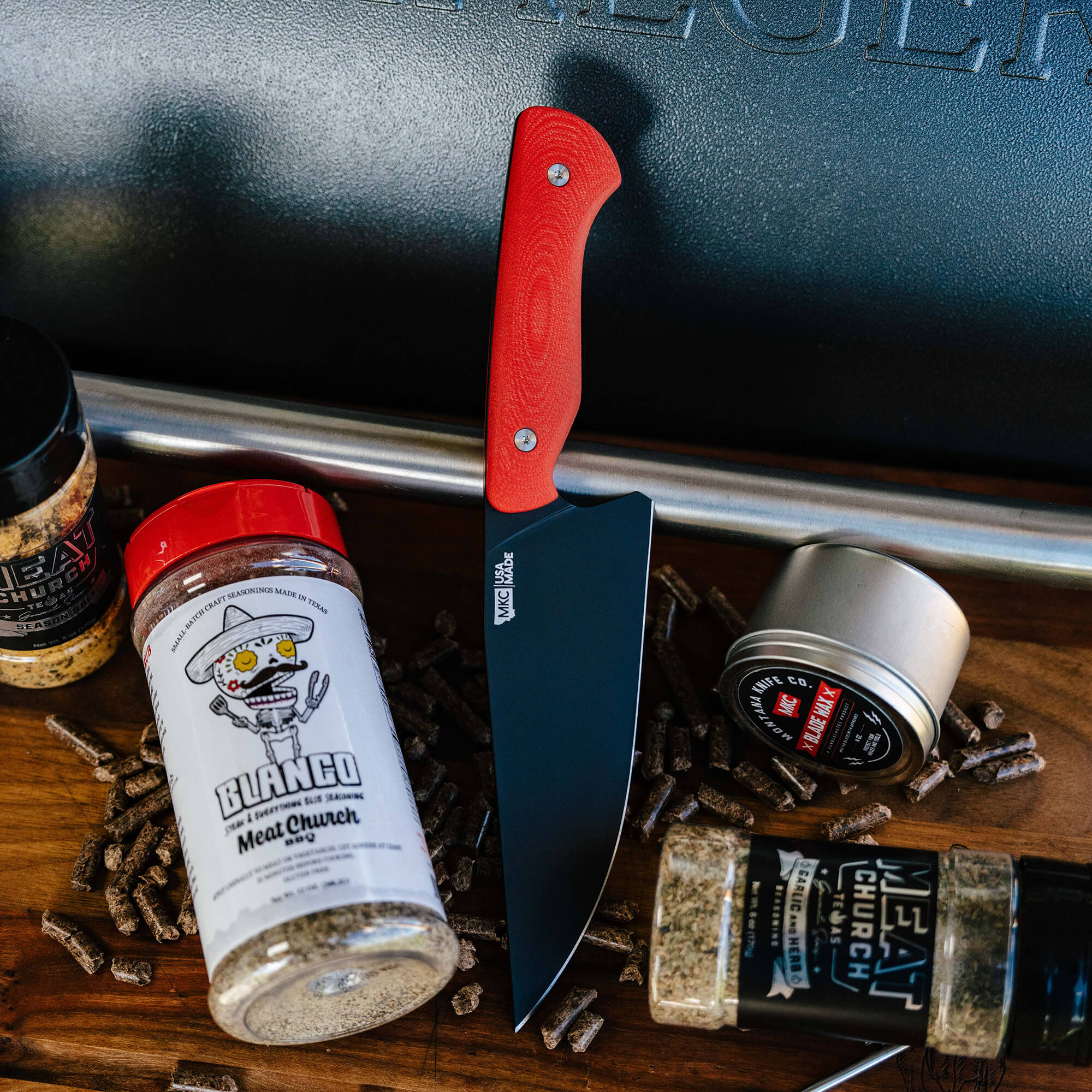 Alt text: "A Meat Church chef knife with a vibrant red handle leaning against a barbecue pit, surrounded by various Meat Church spice rubs including Blanco Meat Church seasoning and Meat Church BBQ rub."