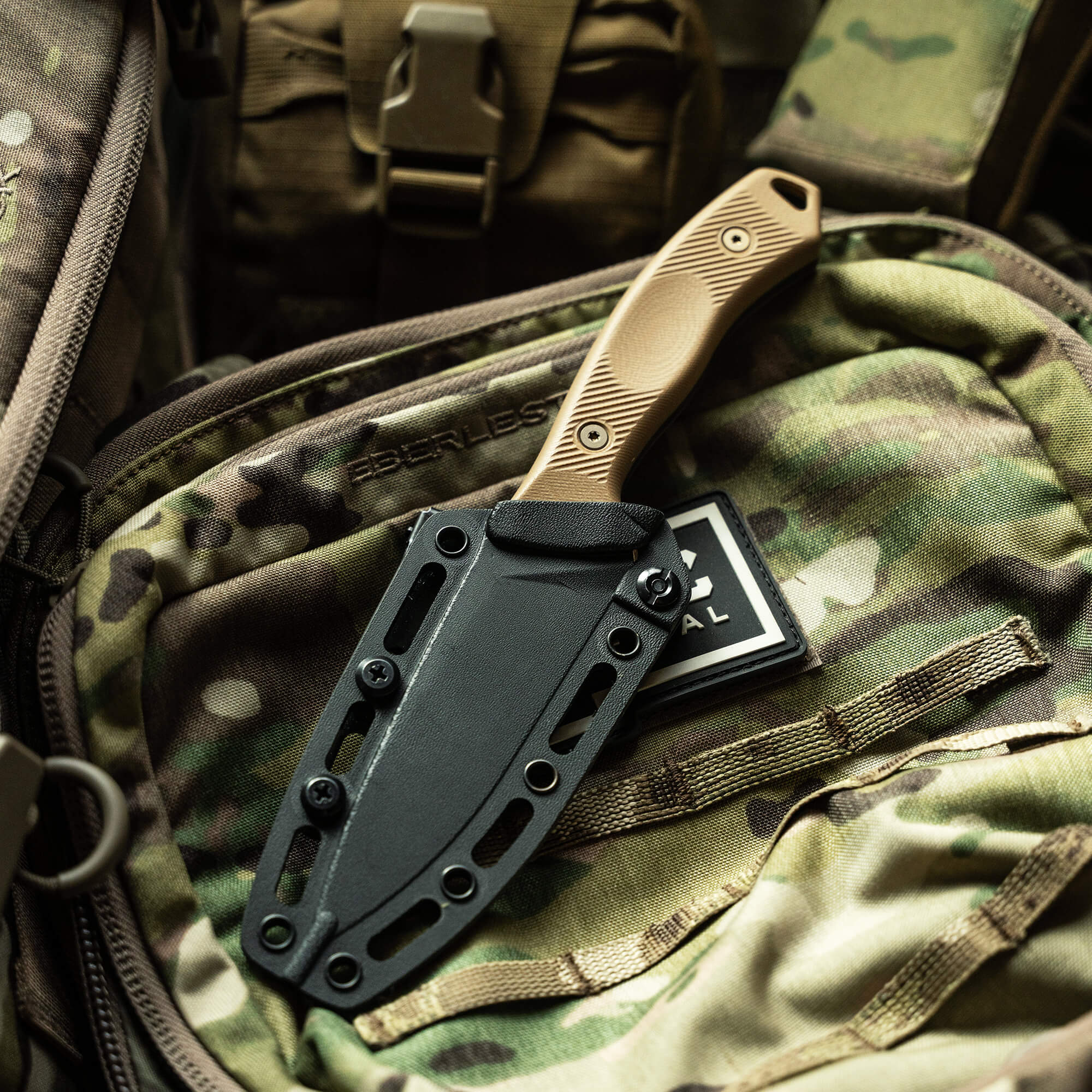 Alt text: "TF24 Kydex sheath in black attached securely to a camouflage backpack, featuring a tan handle knife partially sheathed, emphasizing durability and outdoor utility."