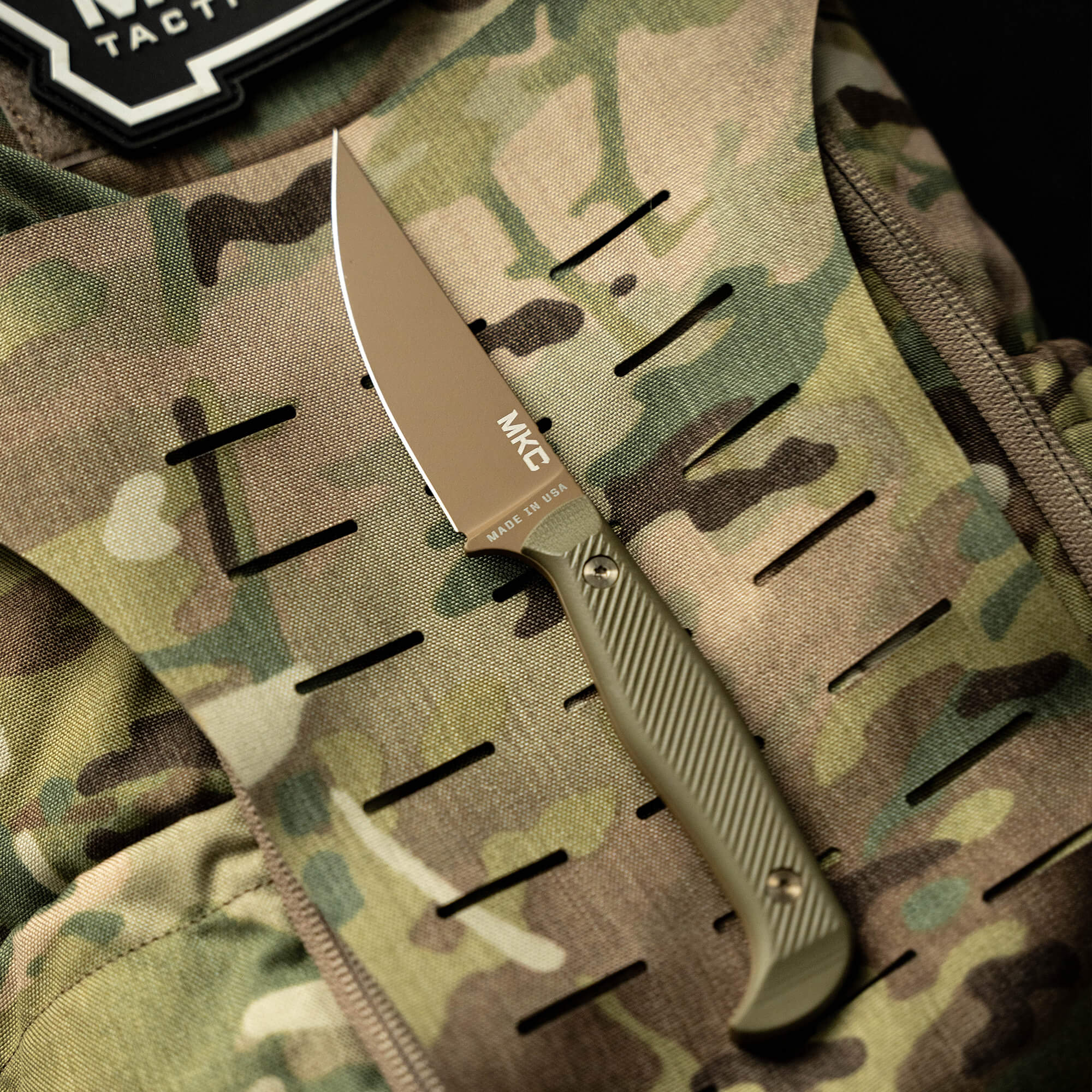 Alt text: "Close-up of a tactical knife, the Battle Goat Coy/OD, with a tan blade marked 'MKE MADE IN USA' on a camouflaged tactical backpack. The knife features a textured olive drab handle."