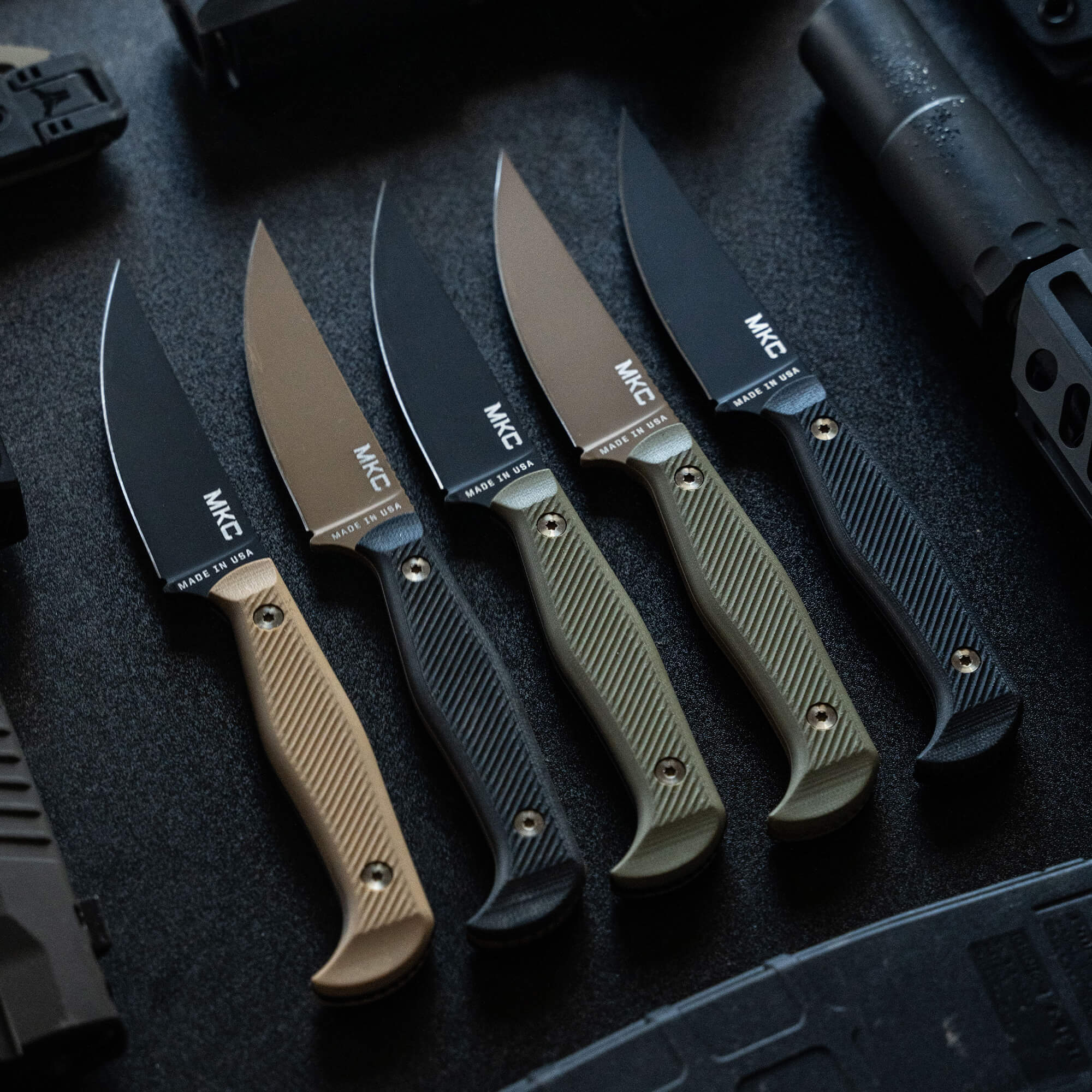 Alt text: "Array of Battle Goat knives in black and olive drab featuring sleek designs with embossed handles, displayed on a tactical surface with gun parts visible on the right."