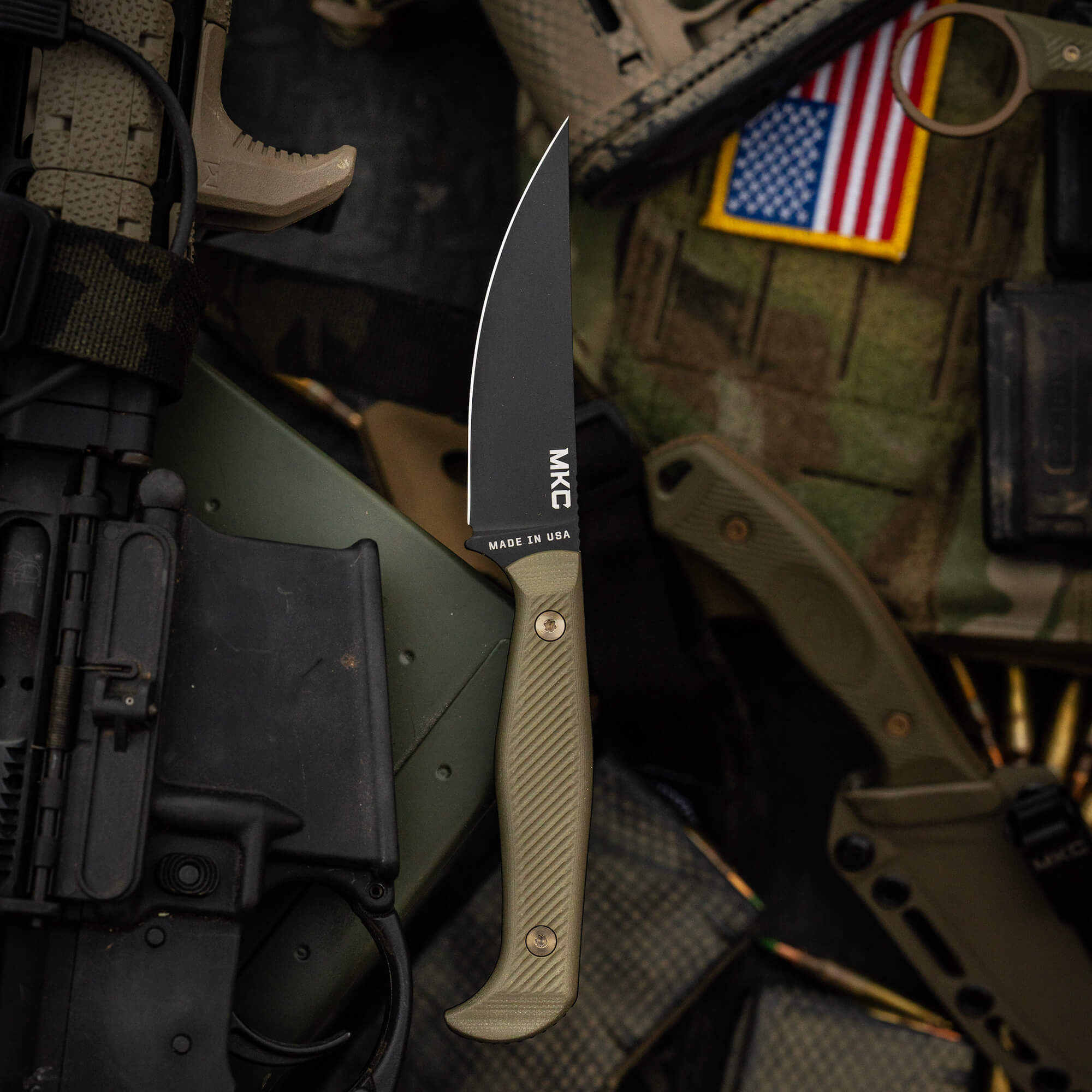 Alt text: "Close-up of the Battle Goat tactical knife in black and olive drab color scheme, featuring a curved blade and textured handle. The knife is prominently displayed with tactical gear and an American flag patch in the background, highlighting its Made in