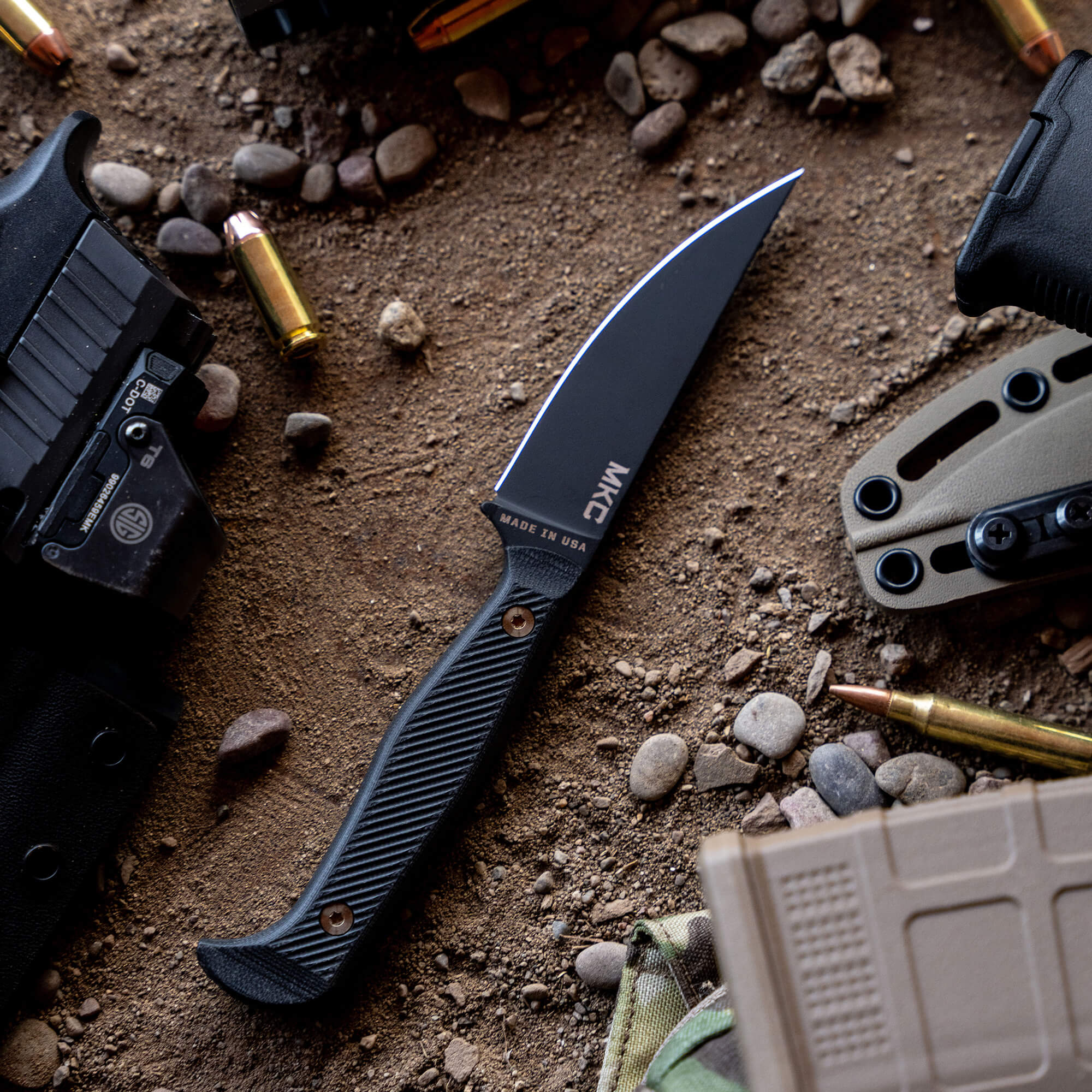 Black tactical fixed-blade knife, marked "Made in USA," lying on a gritty surface with scattered rocks, bullets, a gun holster, and part of a gun visible, emphasizing a rugged, outdoor aesthetic.