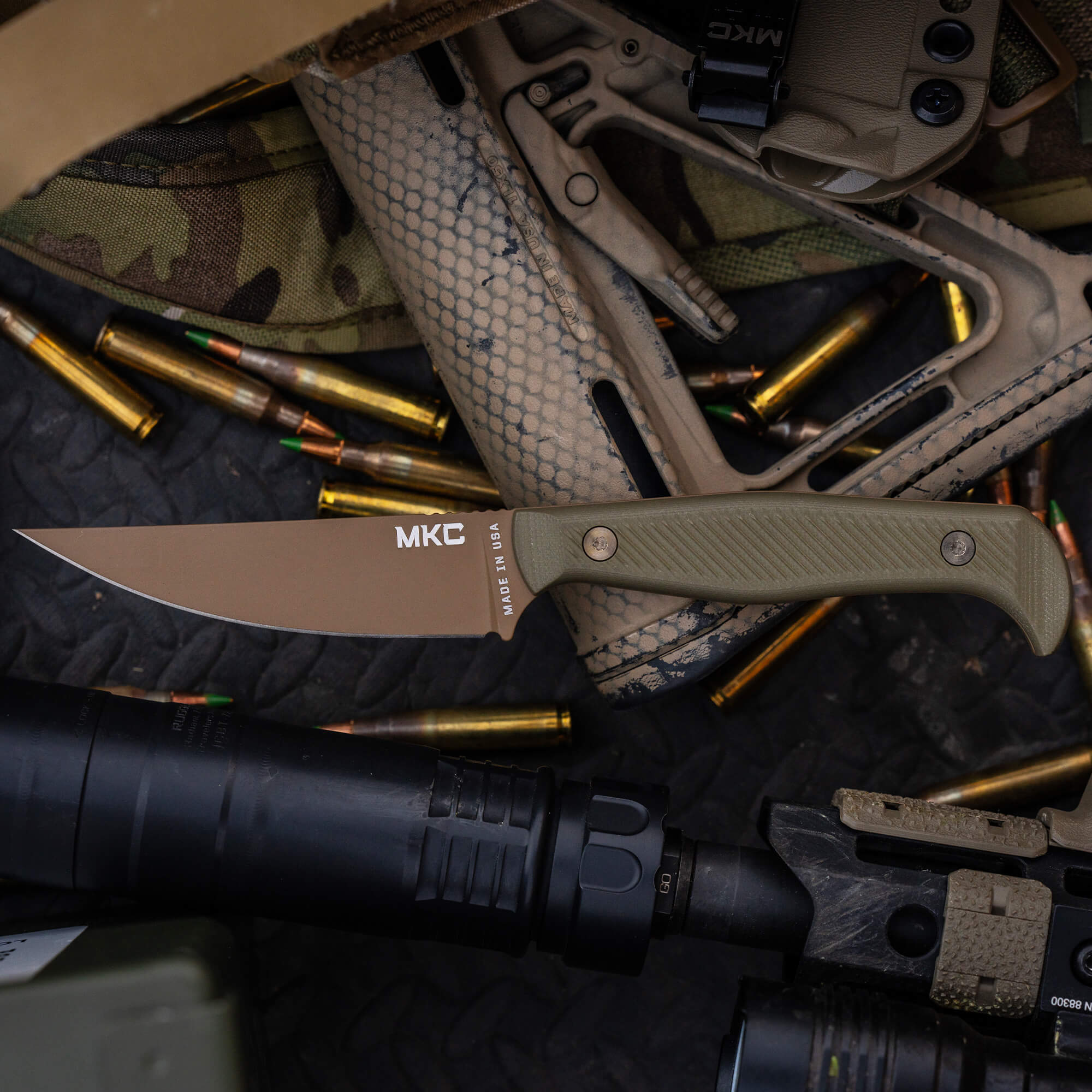 Alt text: "Close-up view of the MKC Battle Goat tactical knife with a tan handle, prominently displayed amid tactical gear including camouflaged fabrics, firearms, and scattered ammunition."