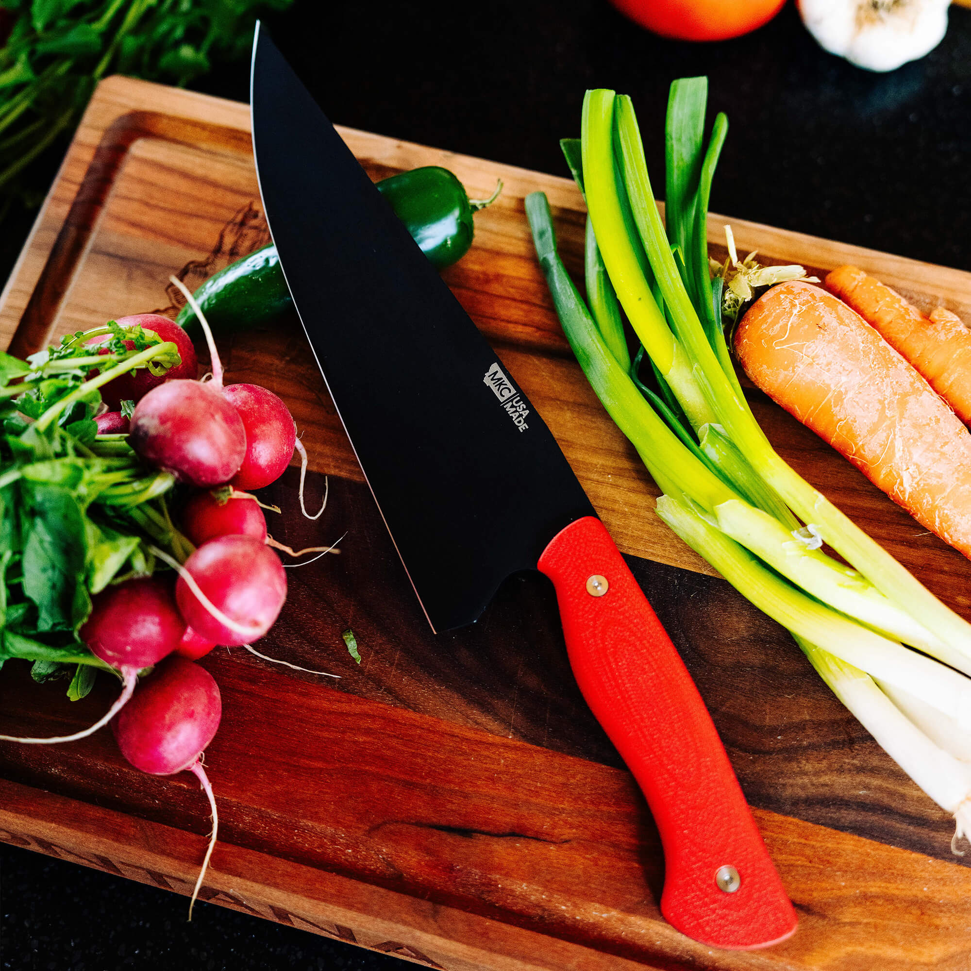 A professional BIGHORN CHEF knife with a red handle lying on a wooden cutting board surrounded by fresh vegetables including radishes, carrots, green peppers, and leeks.