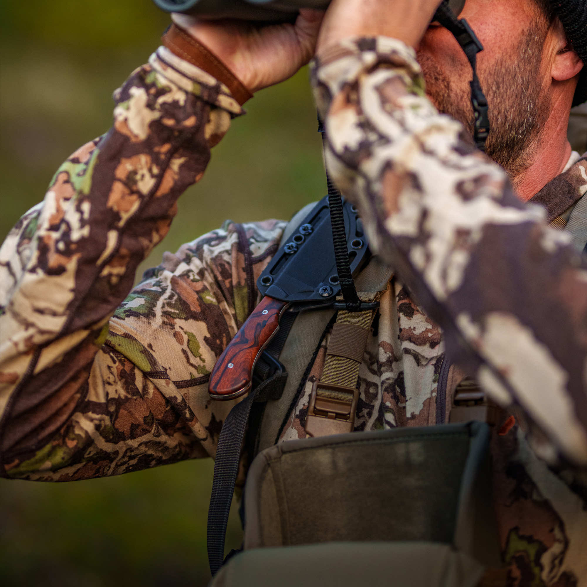 A hunter wearing camouflage gear uses binoculars, showcasing the MKC Elk Knife attached to his gear.