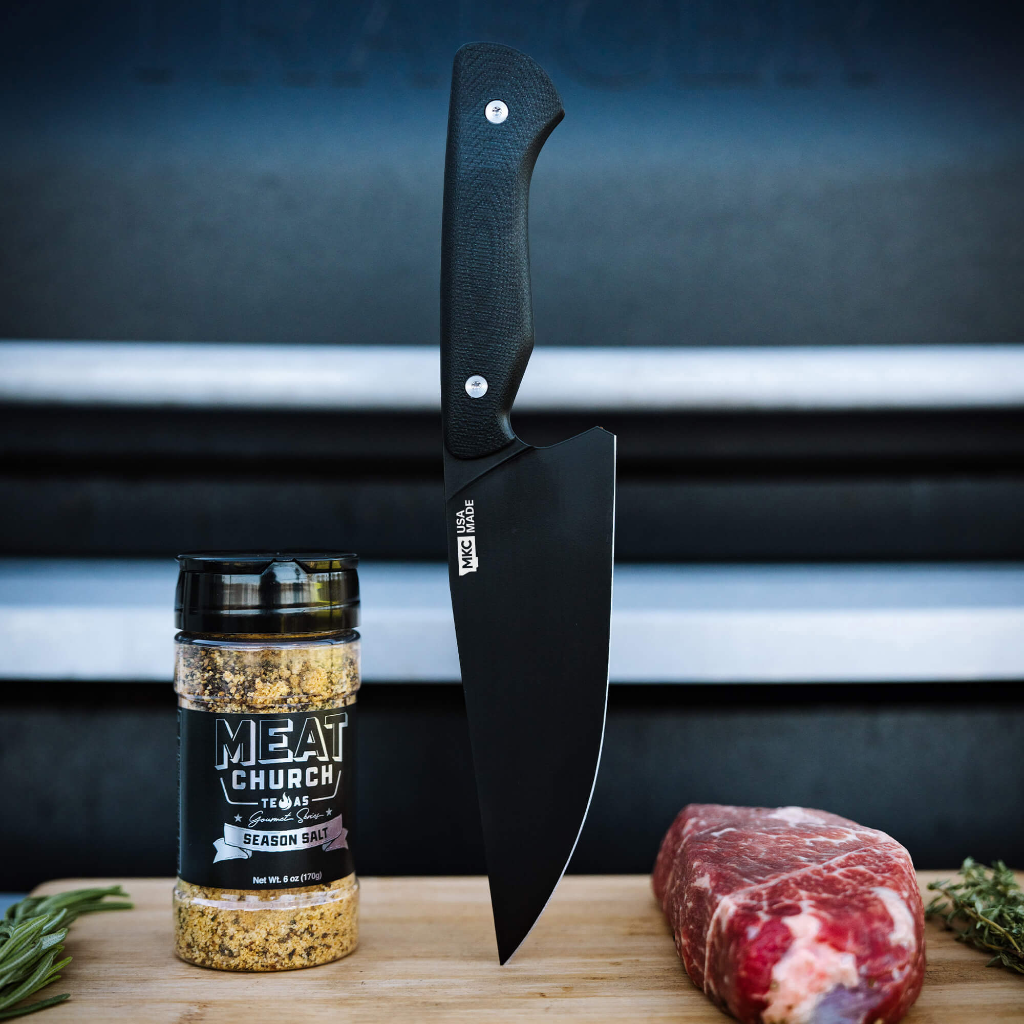 Black Meat Church chef knife displayed alongside seasoning and a cut of steak on a wooden surface.