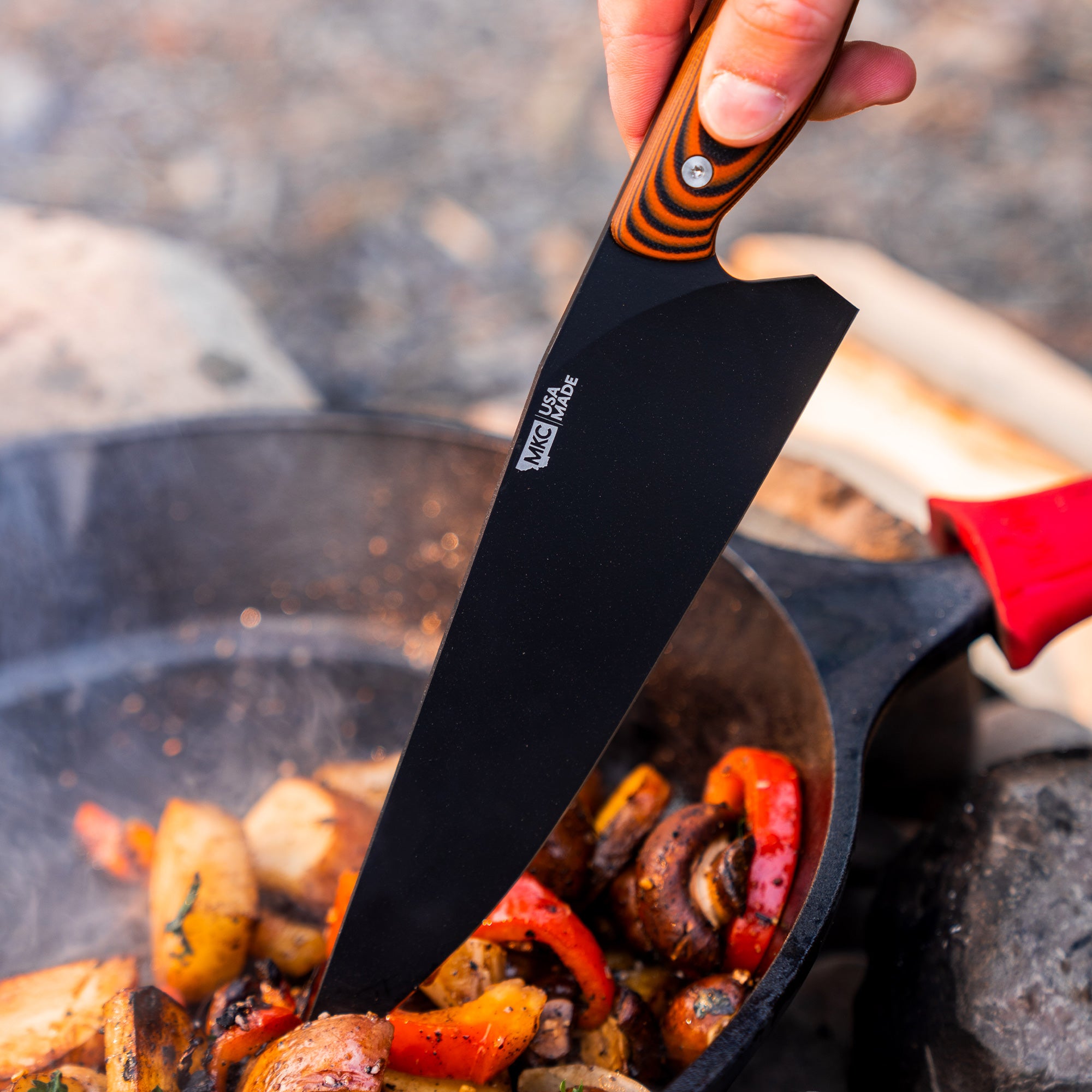 Alt text: "Close-up of a hand holding a BIGHORN Chef knife with orange and black handle, skilfully stirring roasted vegetables in a cast iron skillet over an outdoor campfire."