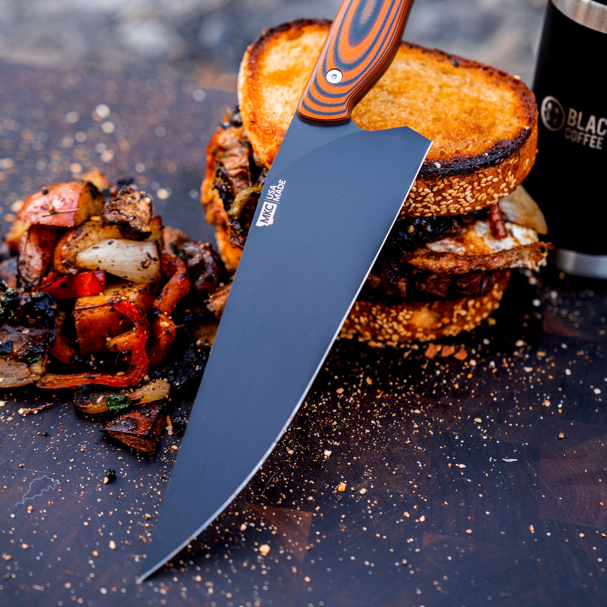 Alt text: "Bighorn Chef orange and black handled chef knife placed on a wooden board next to a sandwich and roasted vegetables, with a Black Coffee thermos cup in the background."