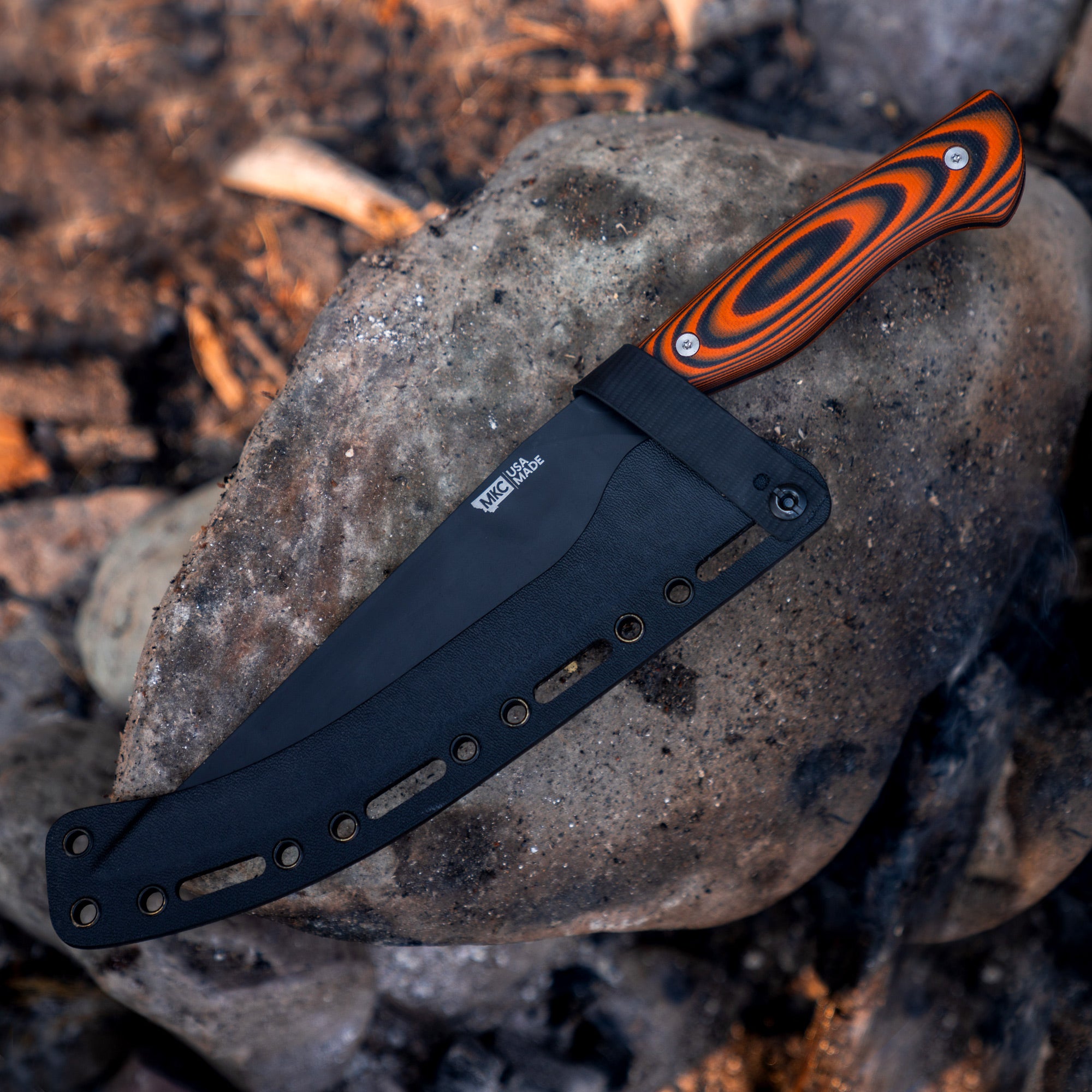 Orange and black Bighorn Chef knife with a textured handle, displayed on a stone surrounded by ashes.