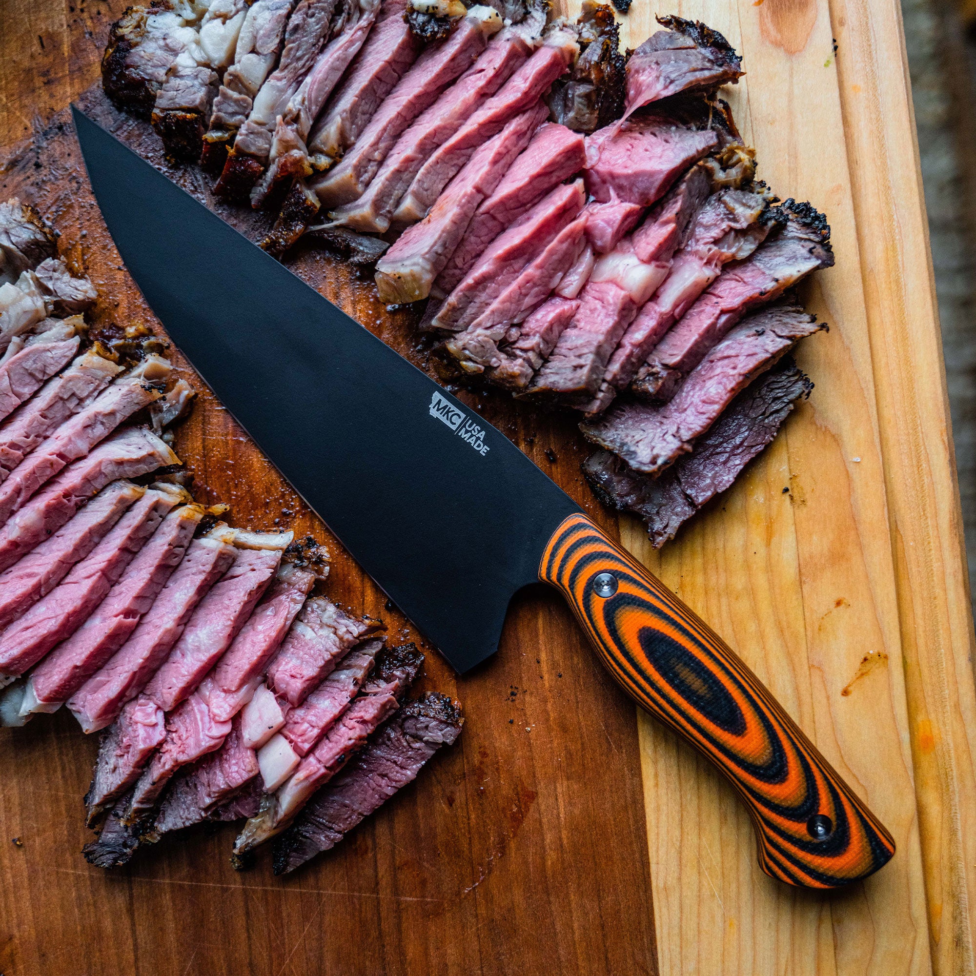 Alt text: "BIGHORN CHEF large chef's knife with an orange and black striped handle, lying beside expertly sliced medium-rare beef brisket on a wooden cutting board."