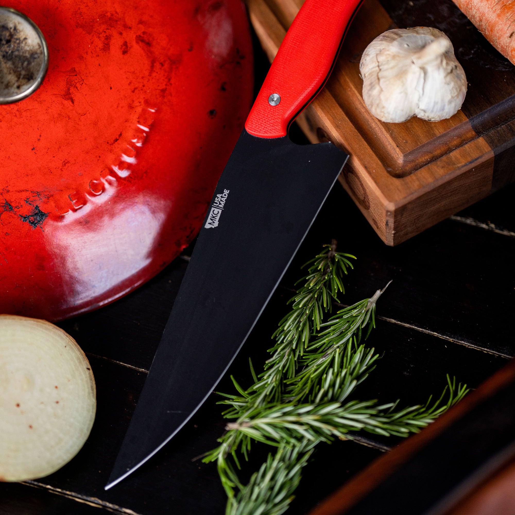 Alt text: "Close-up of a BIGHORN CHEF knife with a red handle, resting on a wooden cutting board next to a red Le Creuset pot lid, garlic, onion, carrot, and fresh rosemary on a dark tiled surface