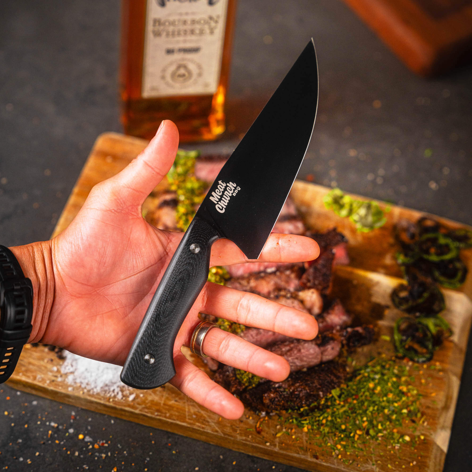Black Meat Church chef knife held in hand, with sliced meat and whiskey bottle in the background on a wooden cutting board.