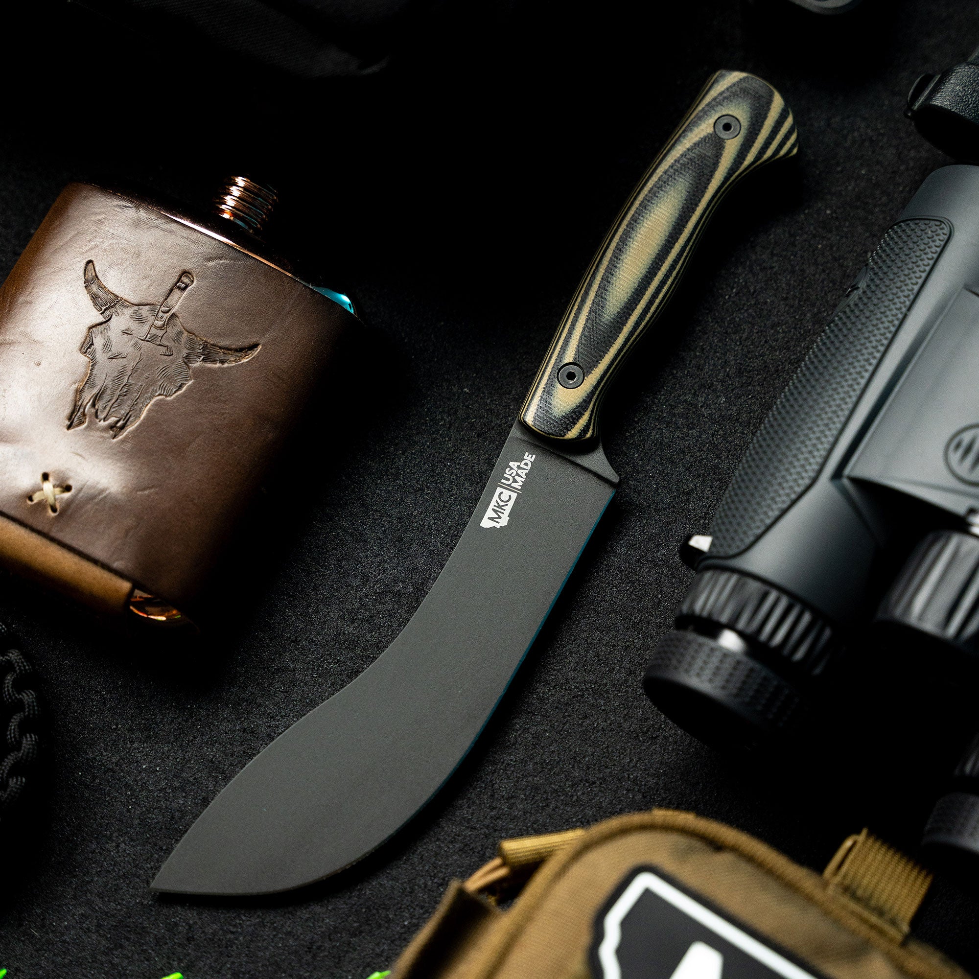 Alt text: "Beartooth Pro Skinning Knife with a curved, black blade and tan and black striped handle, displayed alongside a leather flask and tactical gear on a dark background."