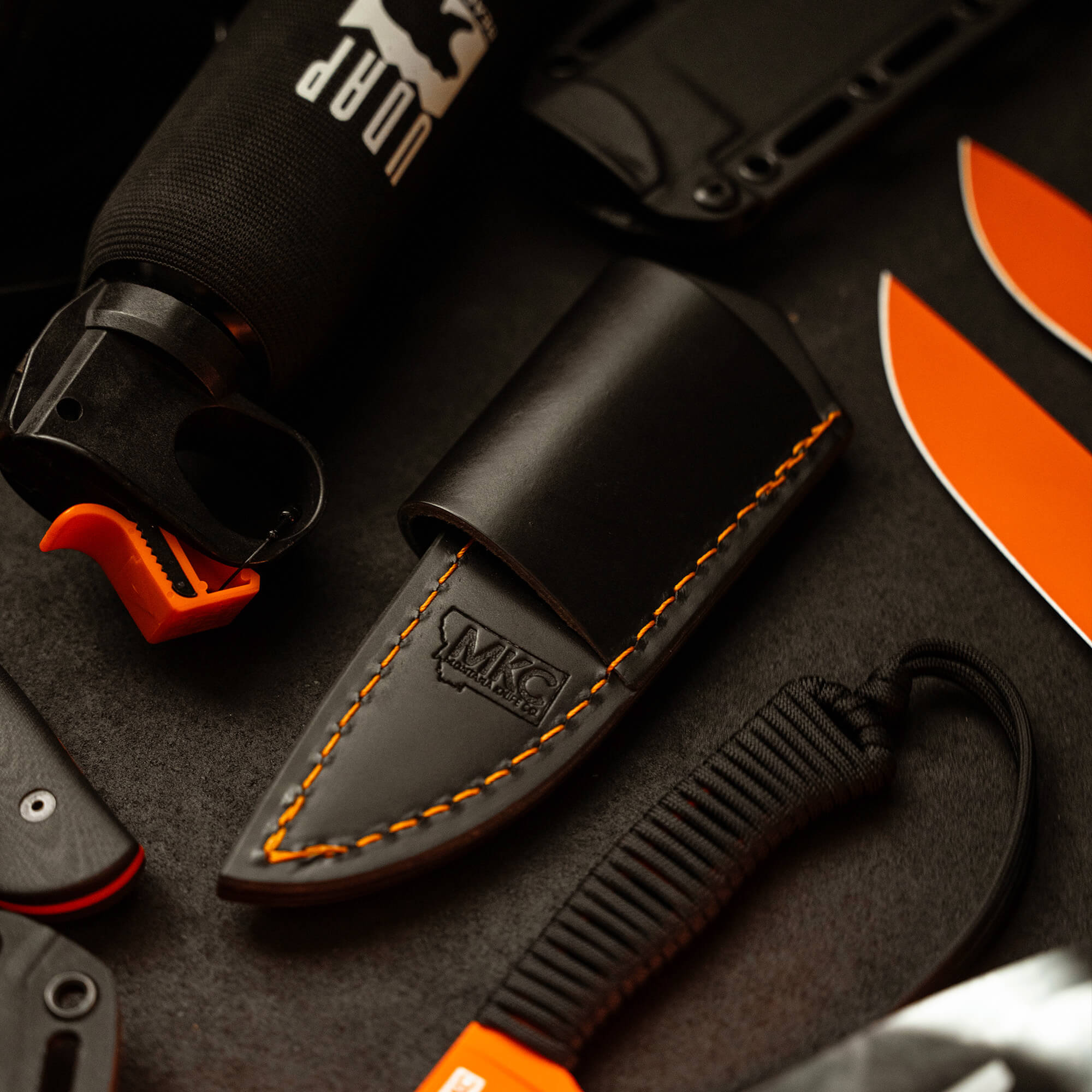 Alt text: Black leather sheath with orange stitching designed for horizontal carry, prominently displaying the MKC logo, surrounded by various outdoor gear including a black water bottle, knife handle, and orange-bladed knife.