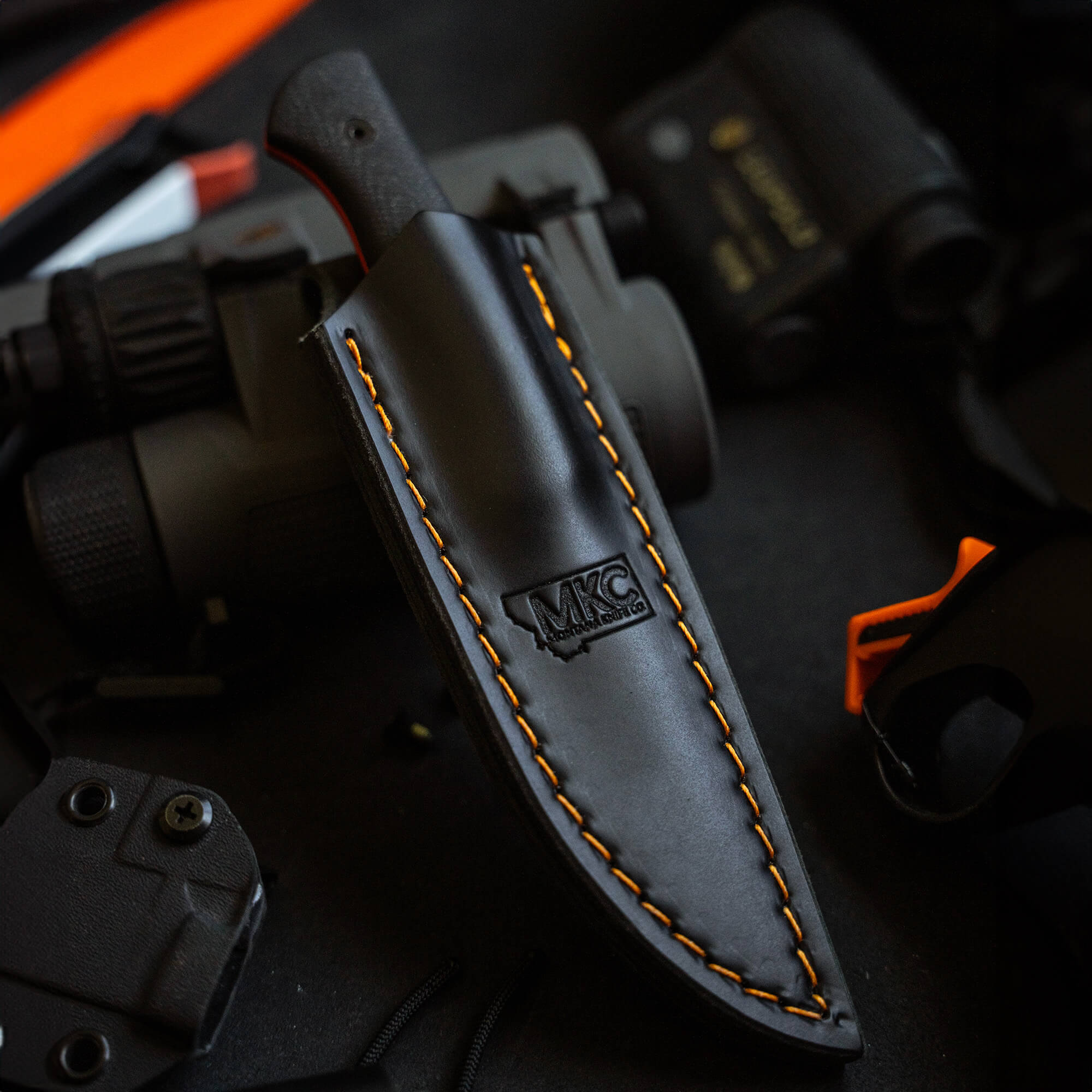 Black leather sheath for vertical carry with orange stitching and MKC logo, set against a dark background with tactical gear.
