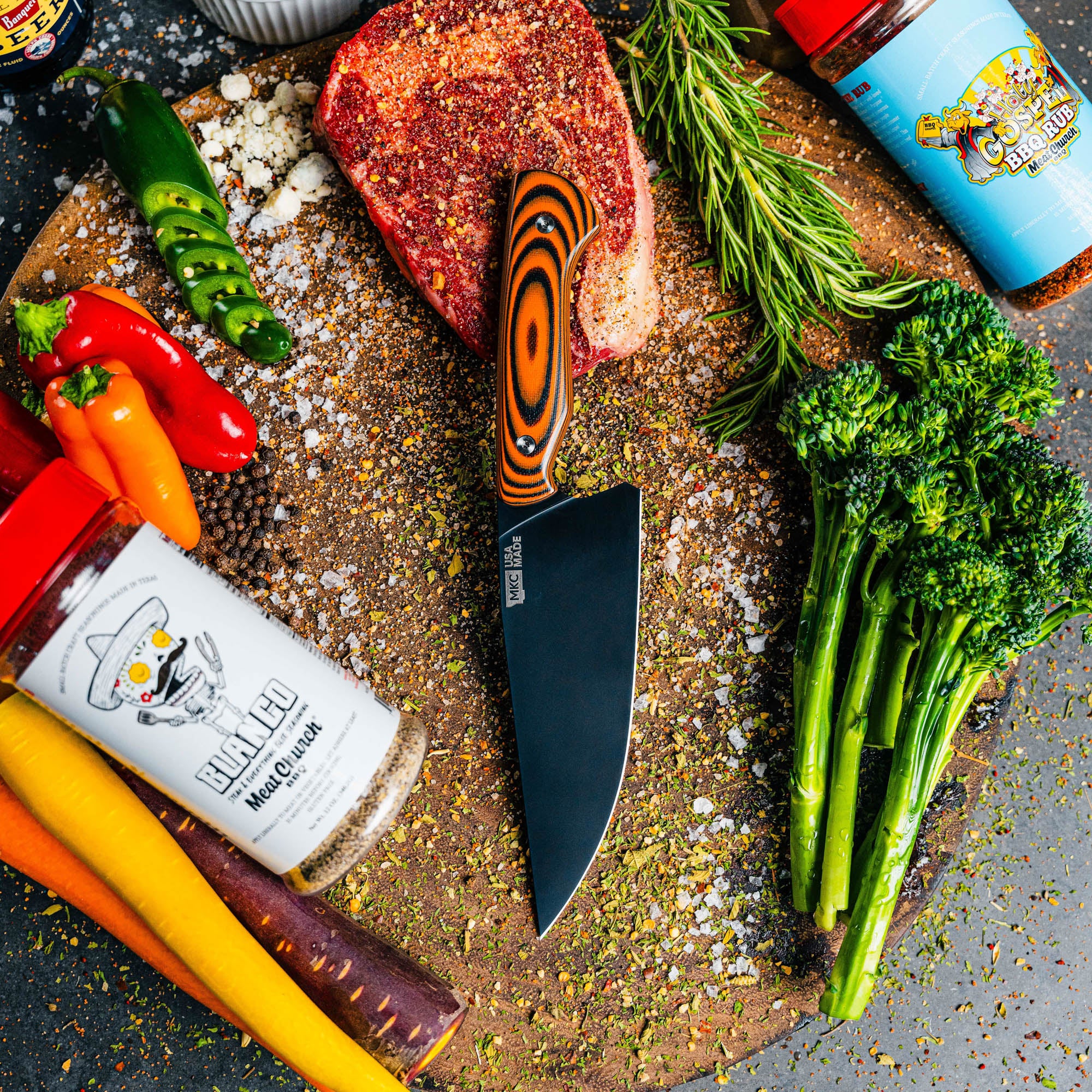 Meat Church chef knife with orange and black handle on a wooden cutting board surrounded by fresh vegetables and spices.