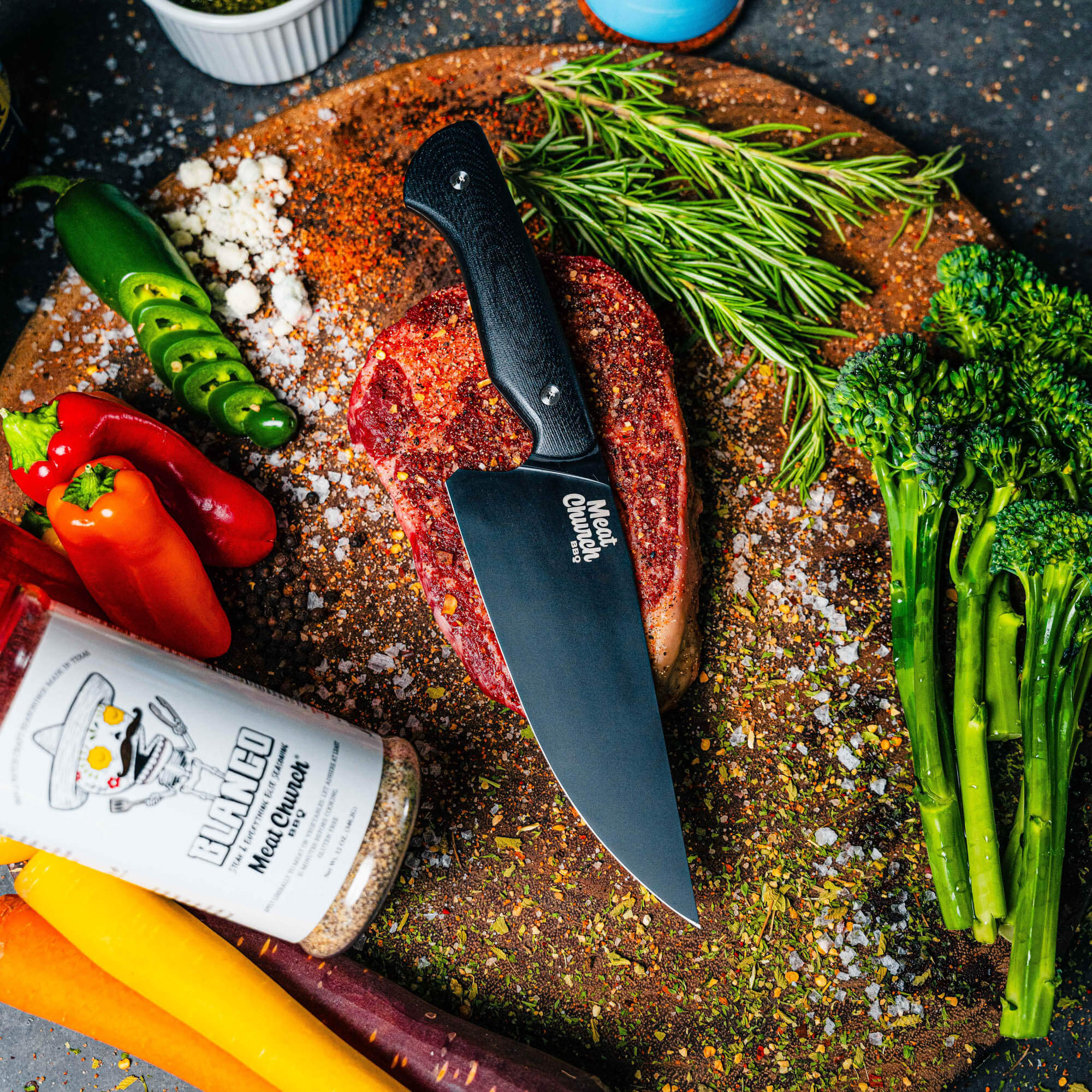 Meat Church chef knife in black on a wooden cutting board with fresh vegetables and seasoned steak, showcasing culinary tools.