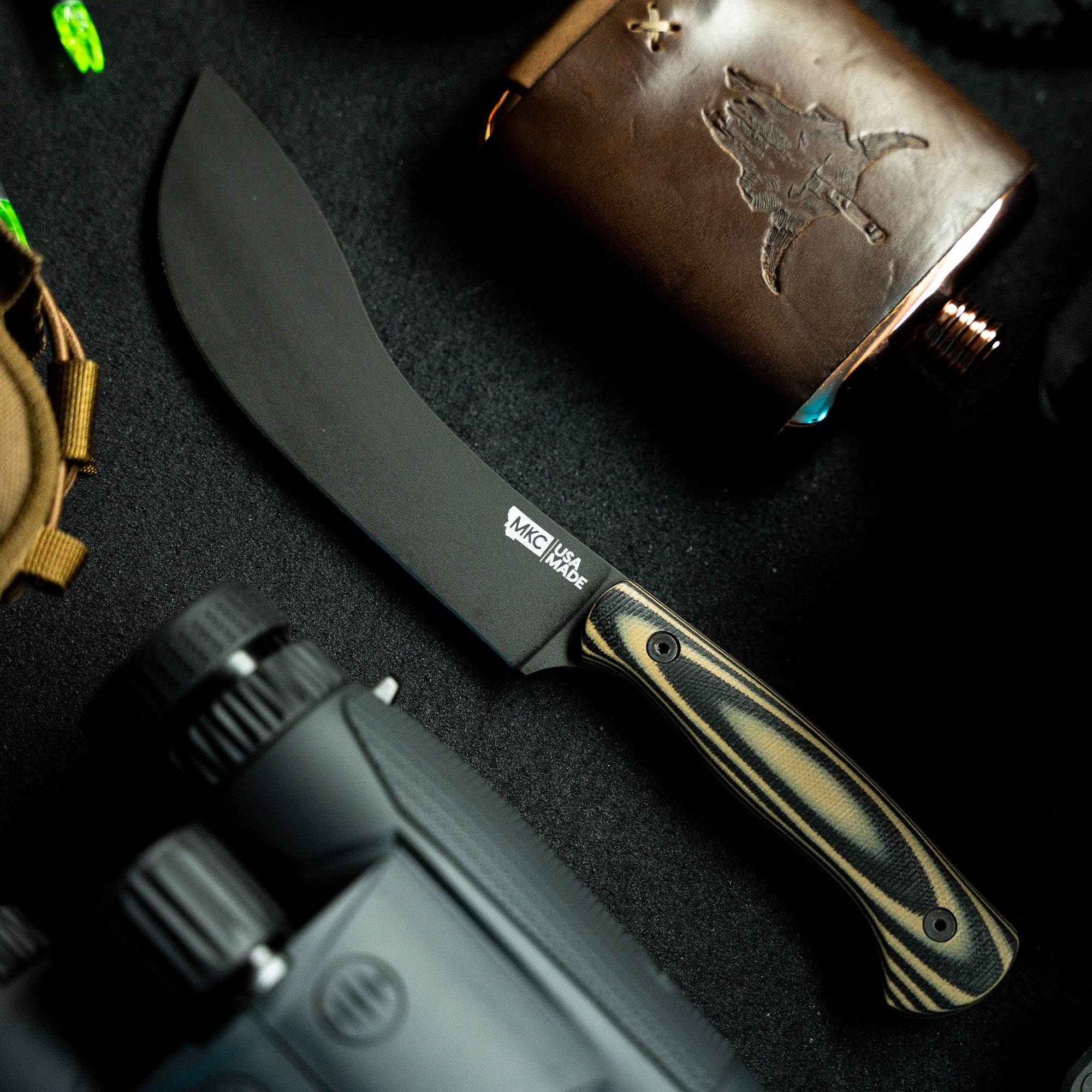 Alt Text: "Close-up view of the Beartooth Pro Skinning Blade with a tan and black handle, featuring a matte black blade, beside accessories including a leather flask with a deer engraving."