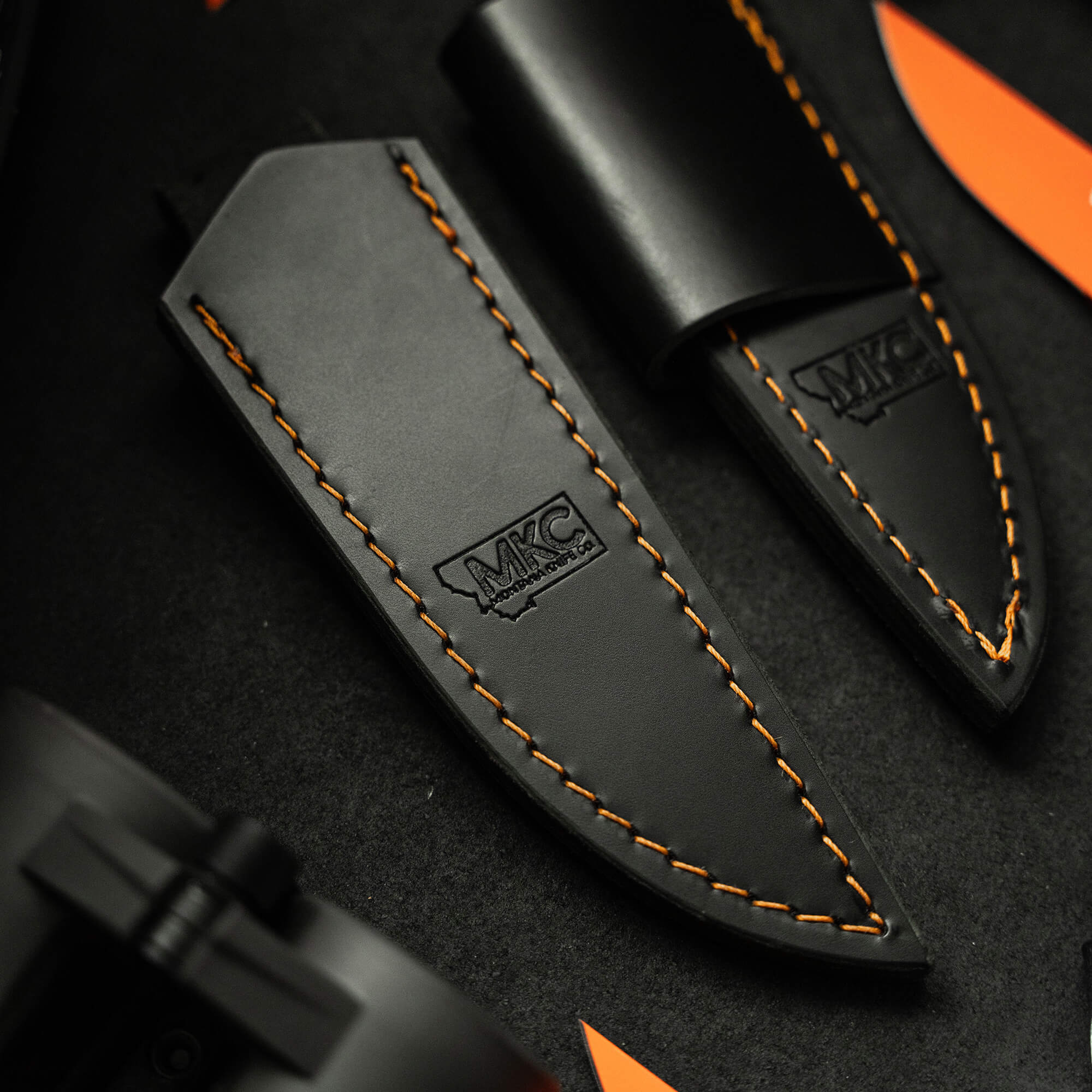 Close-up of a black leather sheath with orange stitching and embossed logo, designed for vertical carry of a knife, displayed on a dark textured background.