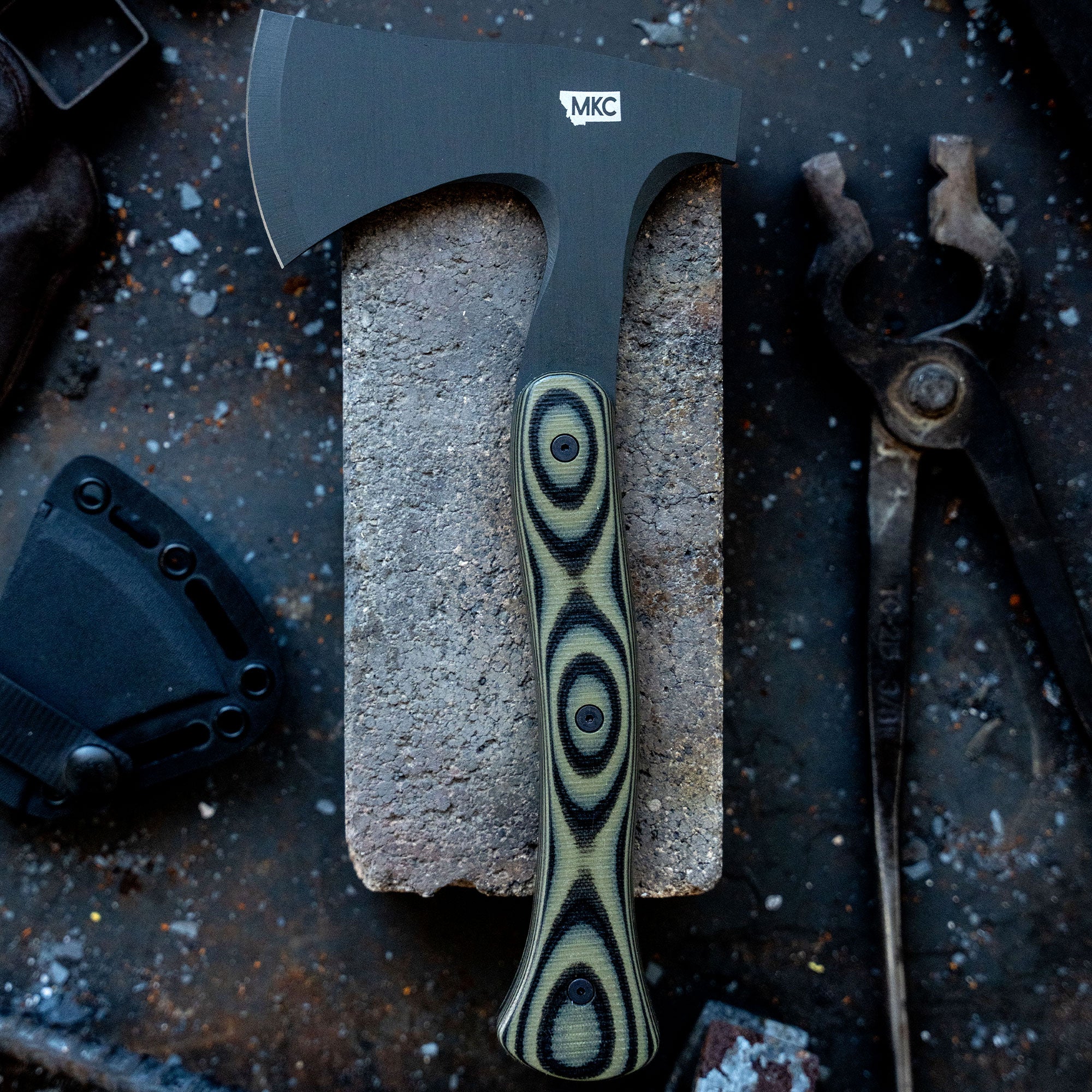 Alt text: "Close-up view of the AEV Hellgate Hatchet with a black blade and micarta handle featuring a distinct circular pattern, displayed on a concrete surface surrounded by industrial tools."