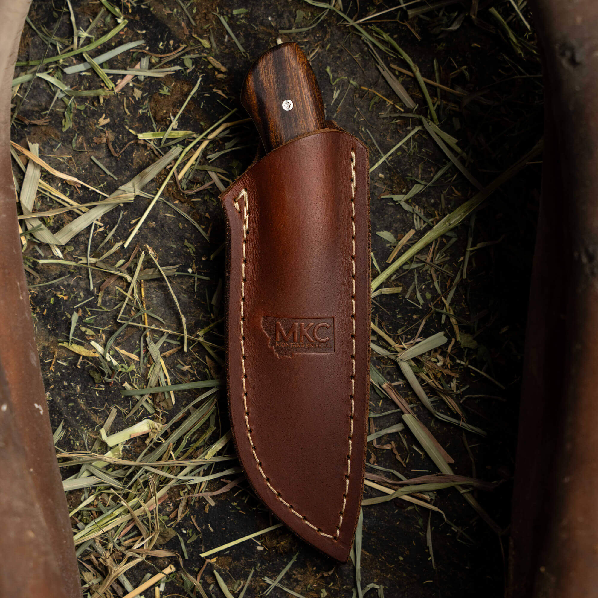 MKC Elk Knife in a brown leather sheath, resting on grass and hay, showcasing craftsmanship for outdoor enthusiasts.