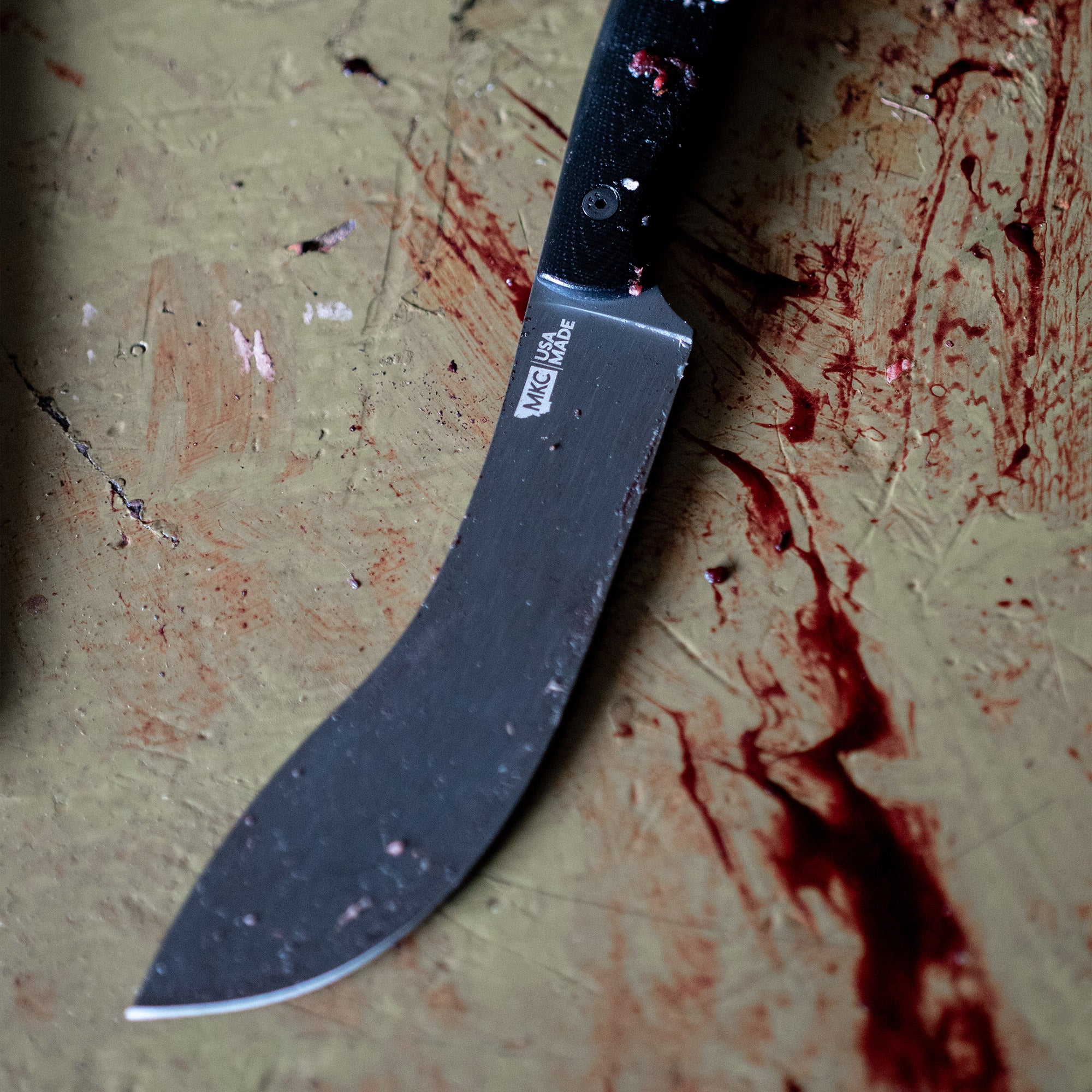Alt text: "Close-up of a Beartooth Pro Skinning Blade with a black handle, resting on a rough surface with visible paint marks and traces of use. The blade is curved and has the 'MNSK' logo near the handle
