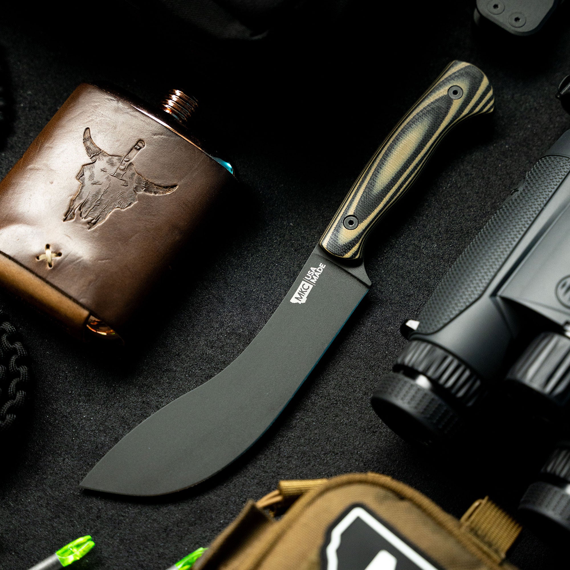 Alt text: "Close-up of the Beartooth Pro Skinning Blade with a tan and black handle, featuring a curved dark matte blade, displayed on a textured surface among various outdoor gear including a leather flask with an embossed design."