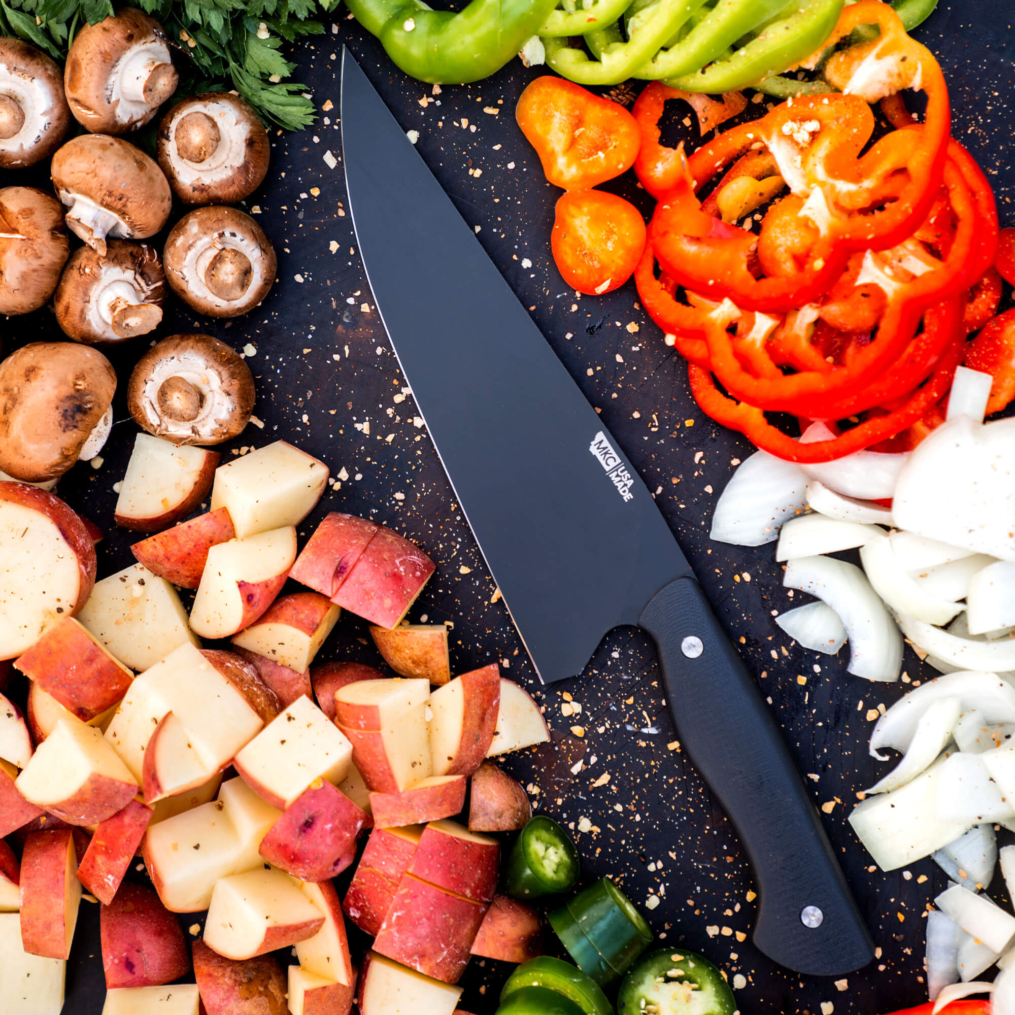 Alt text: "Bighorn Chef black chef's knife displayed on a dark cutting board surrounded by colorful chopped vegetables including red peppers, mushrooms, apples, and onions, with seasonings scattered around, highlighting the knife's sleek design and sharp blade."
