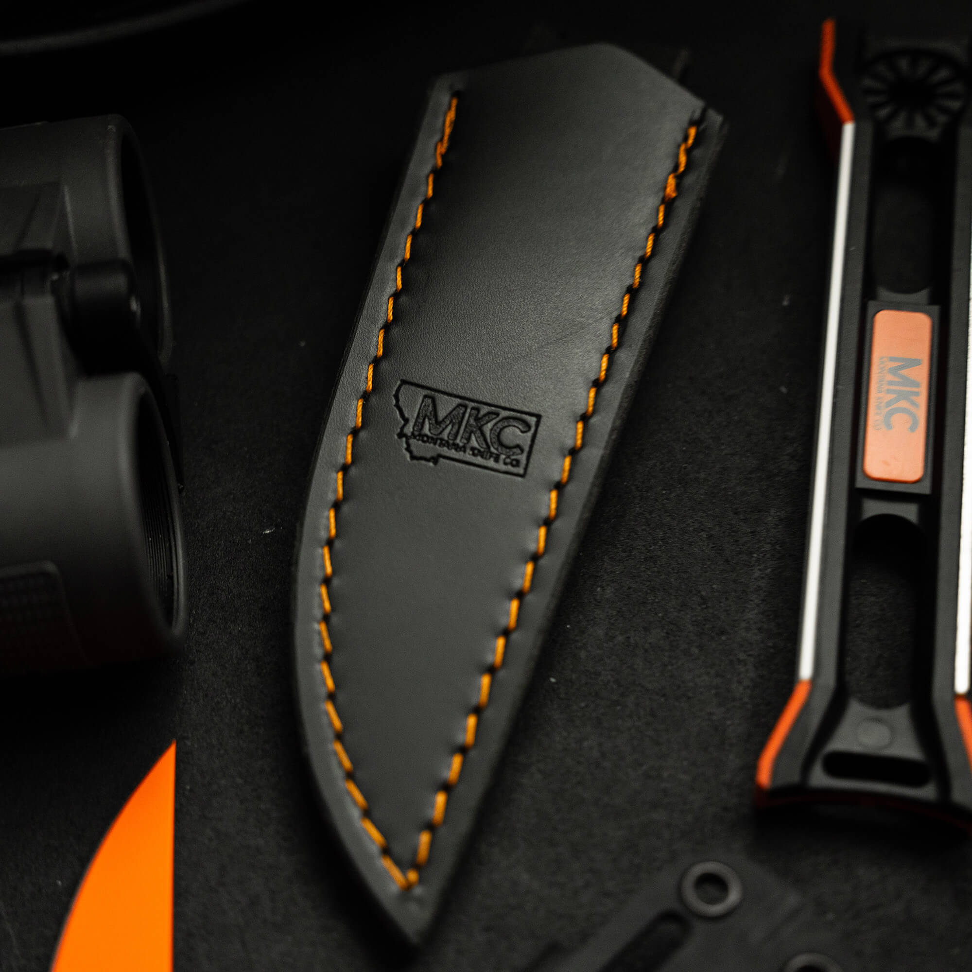 Black and orange Blackfoot 2.0 leather sheath for vertical carry, featuring contrast stitching and logo imprint, positioned next to tactical equipment on a dark surface.