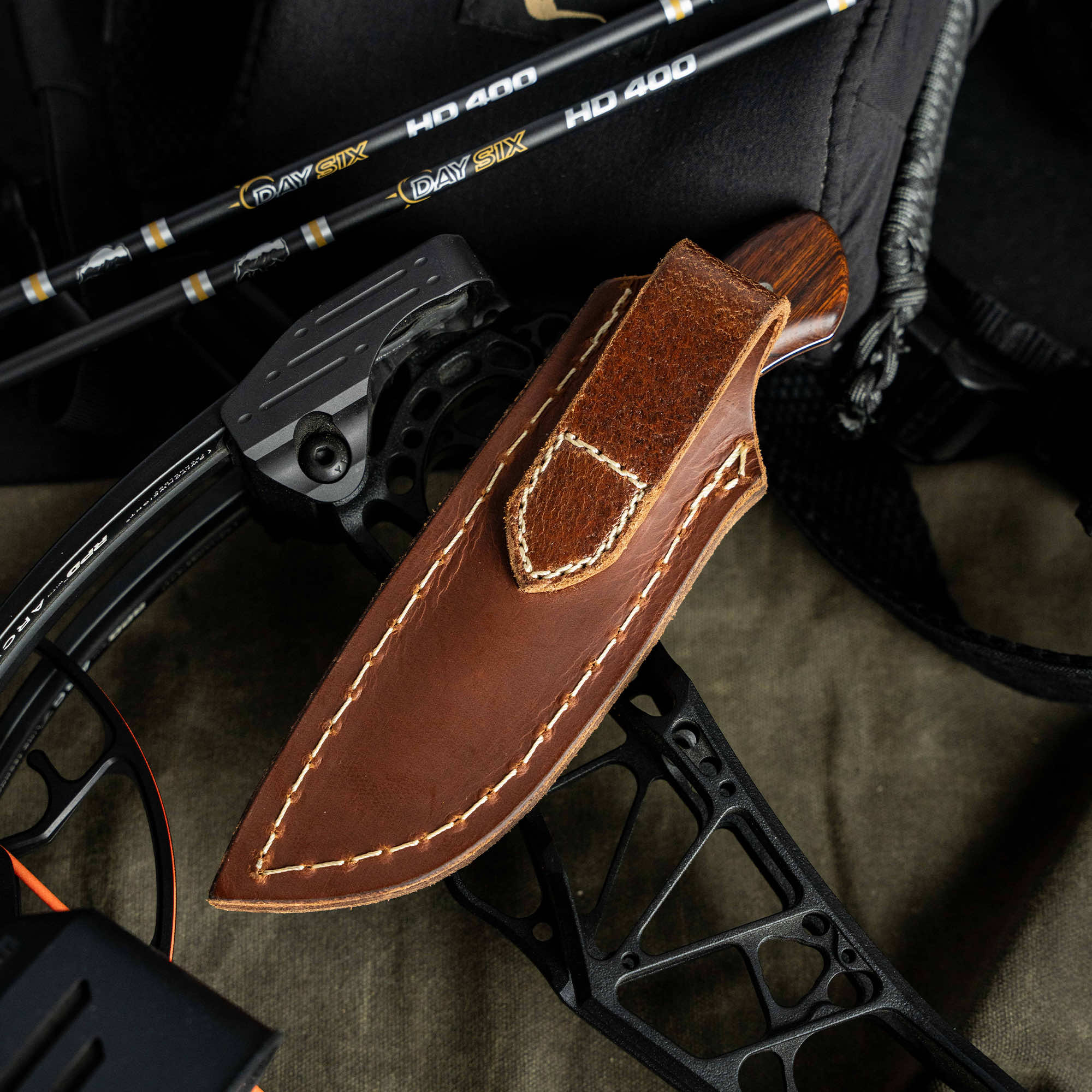 Montana Knife Company's LIVE WILD Traditions Elk Knife in leather sheath, resting on a bow and arrows, ideal for outdoor enthusiasts.