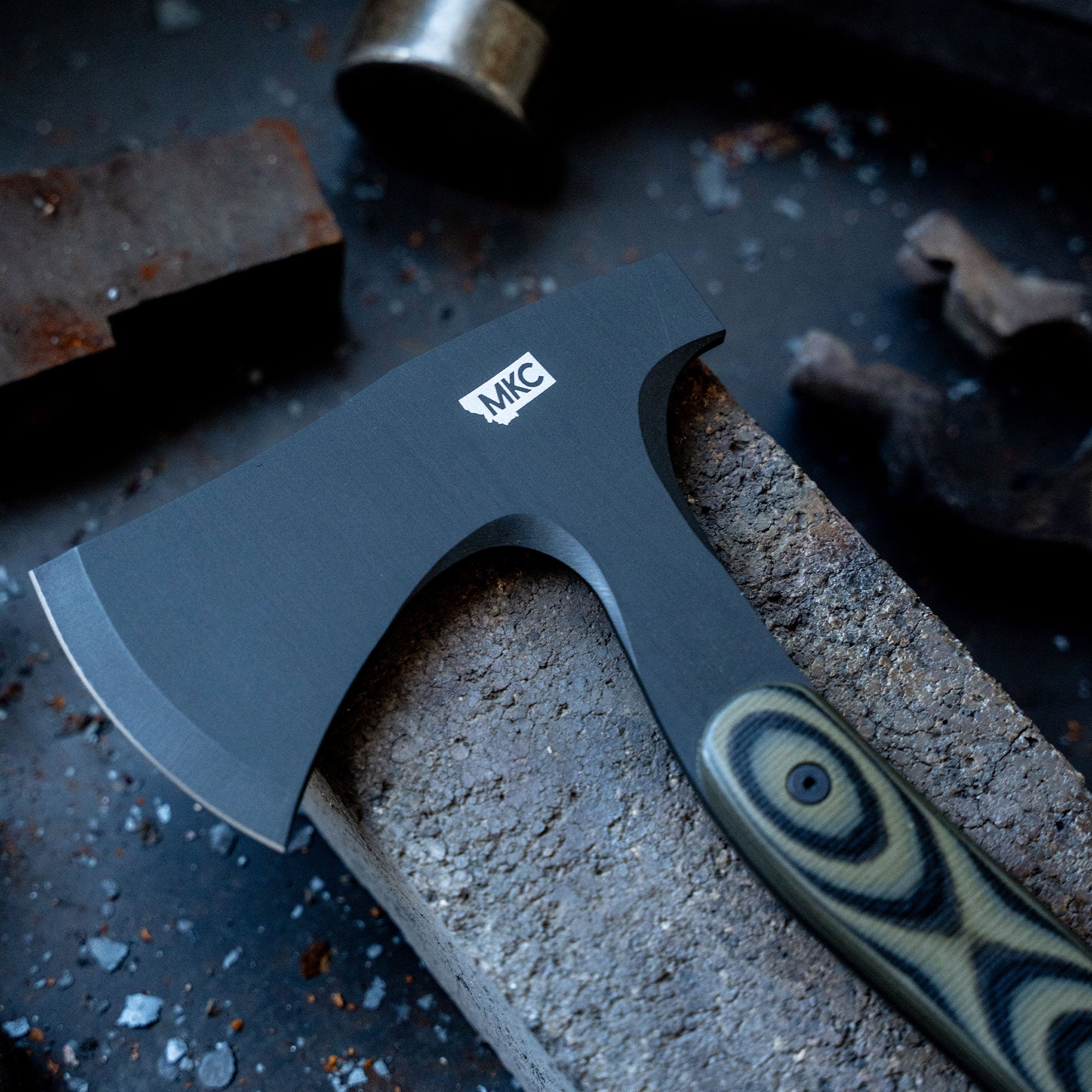 Alt text: "Close-up of the AEV Hellgate Hatchet with MKC logo, placed on a gritty workshop surface surrounded by tools and metal scraps."