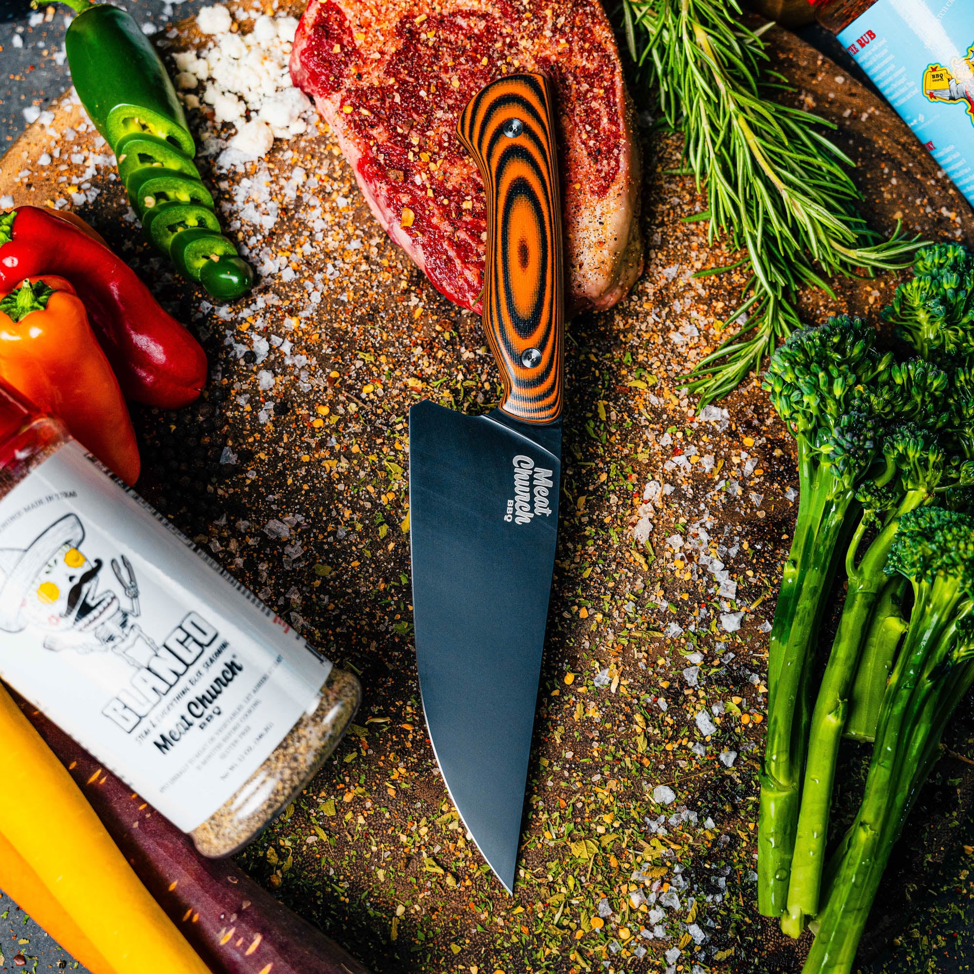 Meat Church chef knife in orange and black on a cutting board surrounded by fresh vegetables and seasonings.