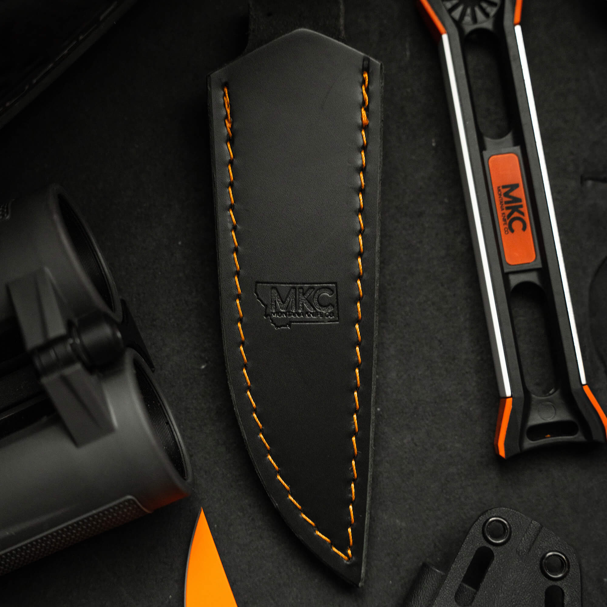 Alt text: Close-up of a black leather sheath with orange stitching, marked with the MKC logo, for vertical carry, displayed on a dark grey background with partial views of knives and outdoor equipment.