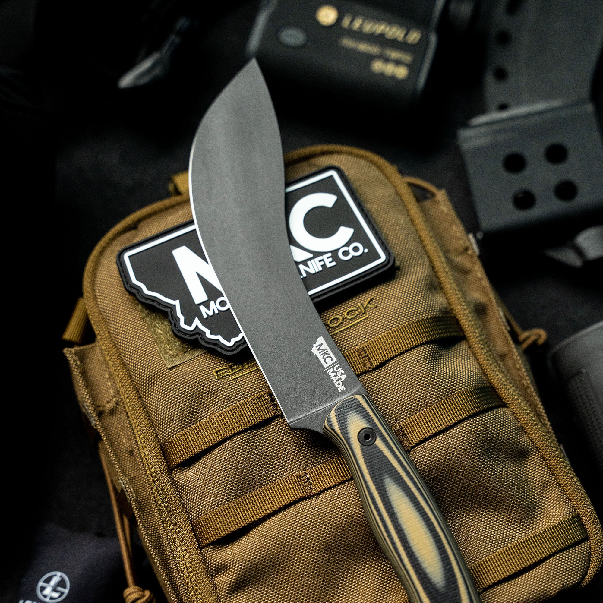 Alt text: Close-up of a Beartooth Pro Skinning knife with a tan and black handle positioned on a tactical backpack. The blade is prominently displayed with the logo 'MKC USA' etched near the handle.