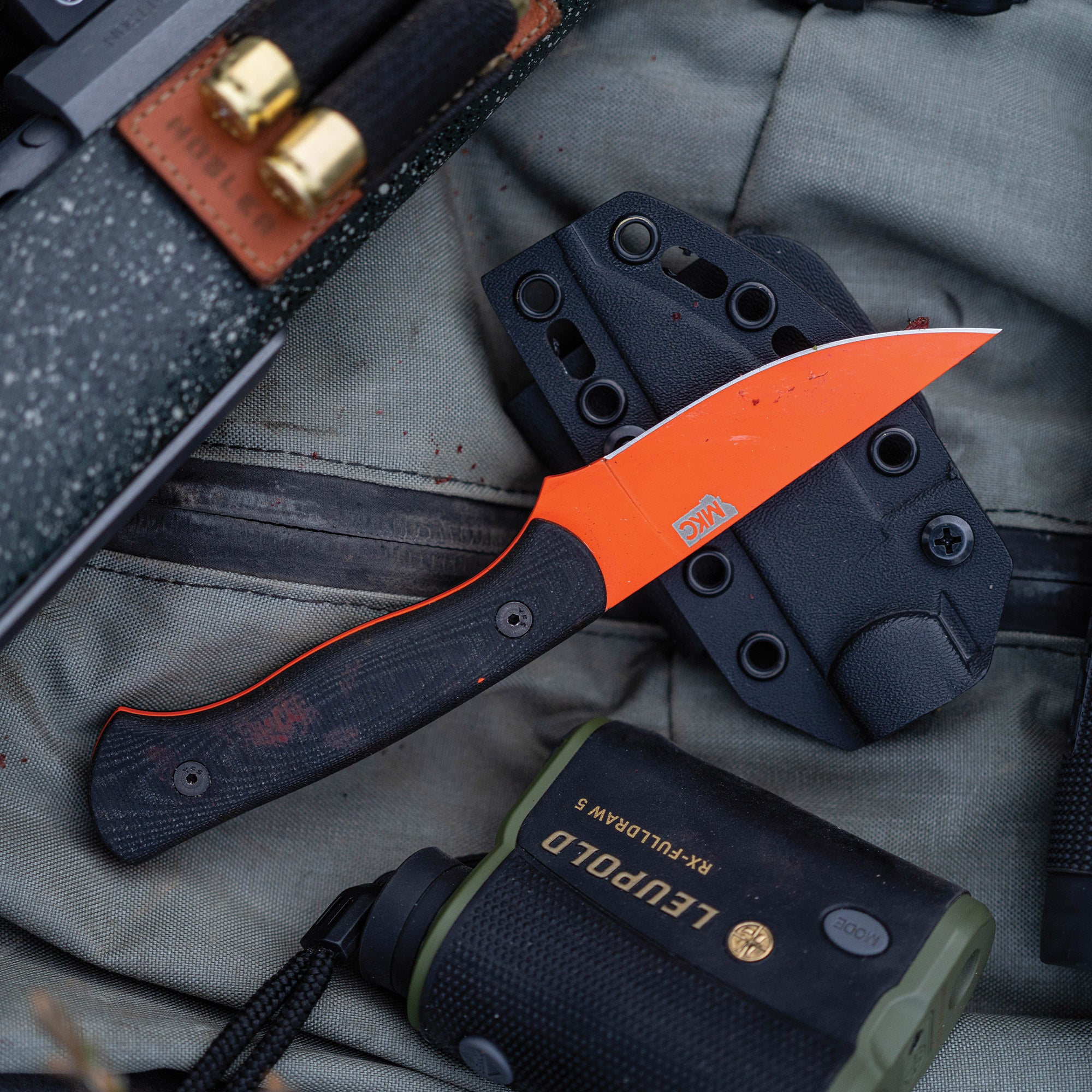 Alt text: "Close-up of the BLACKFOOT 2.0 fixed blade knife with a blaze orange Cerakote™ finish, displayed on a textured grey backpack next to a binocular case and bullets in a leather ammo holder."