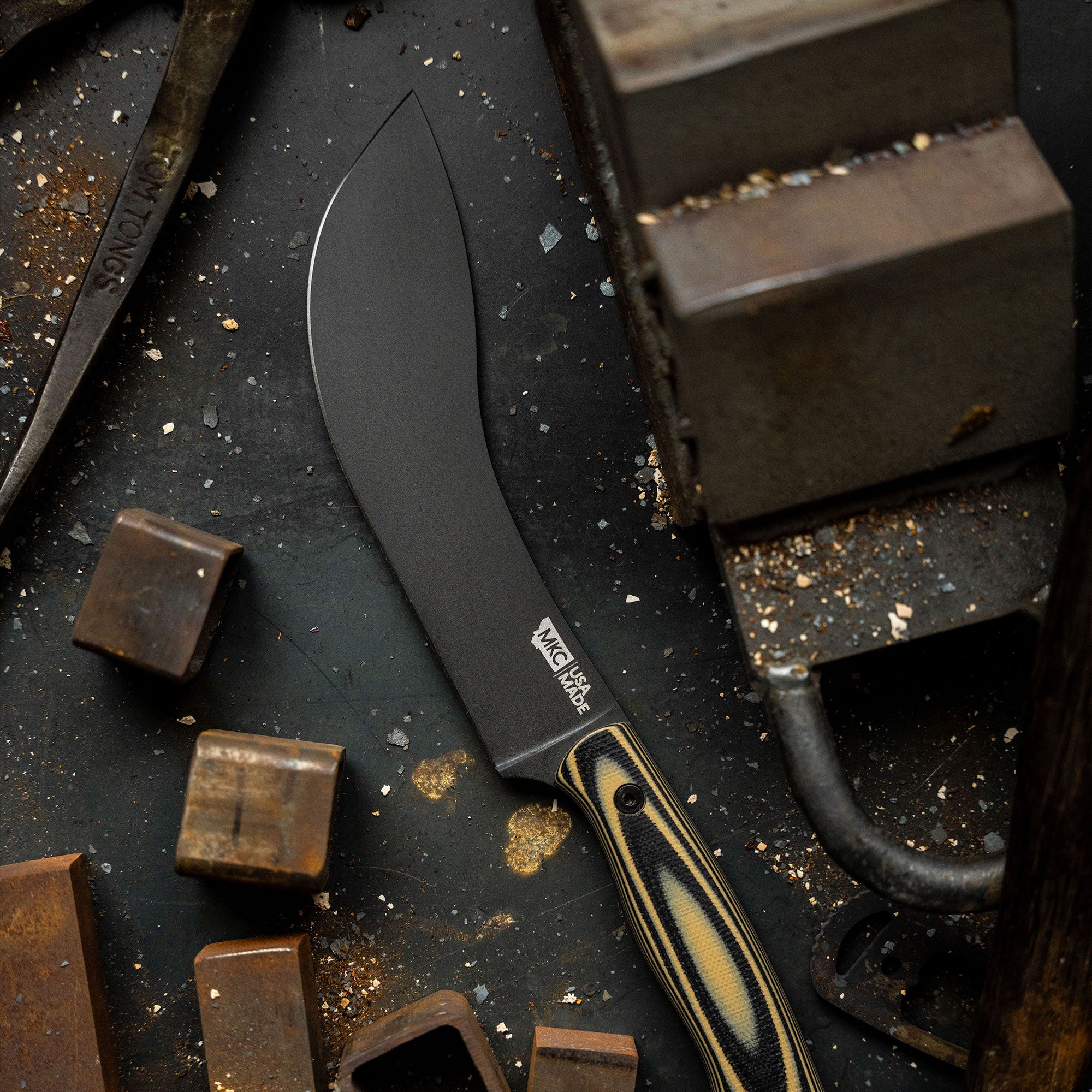 Alt text: "Close-up of the Beartooth Pro Skinning Blade in tan and black, featuring a sharp, broad blade and striped handle, displayed on a rustic dark metal background with scattered metalworking tools and debris."