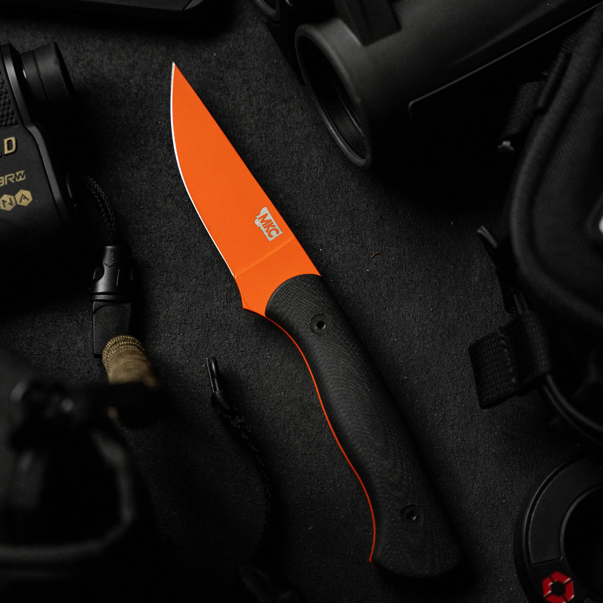 Alt text: "BLACKFOOT 2.0 fixed blade knife with a vibrant blaze orange Cerakote™ blade and black textured handle, displayed amidst tactical gear including a flashlight and binoculars on a dark background."