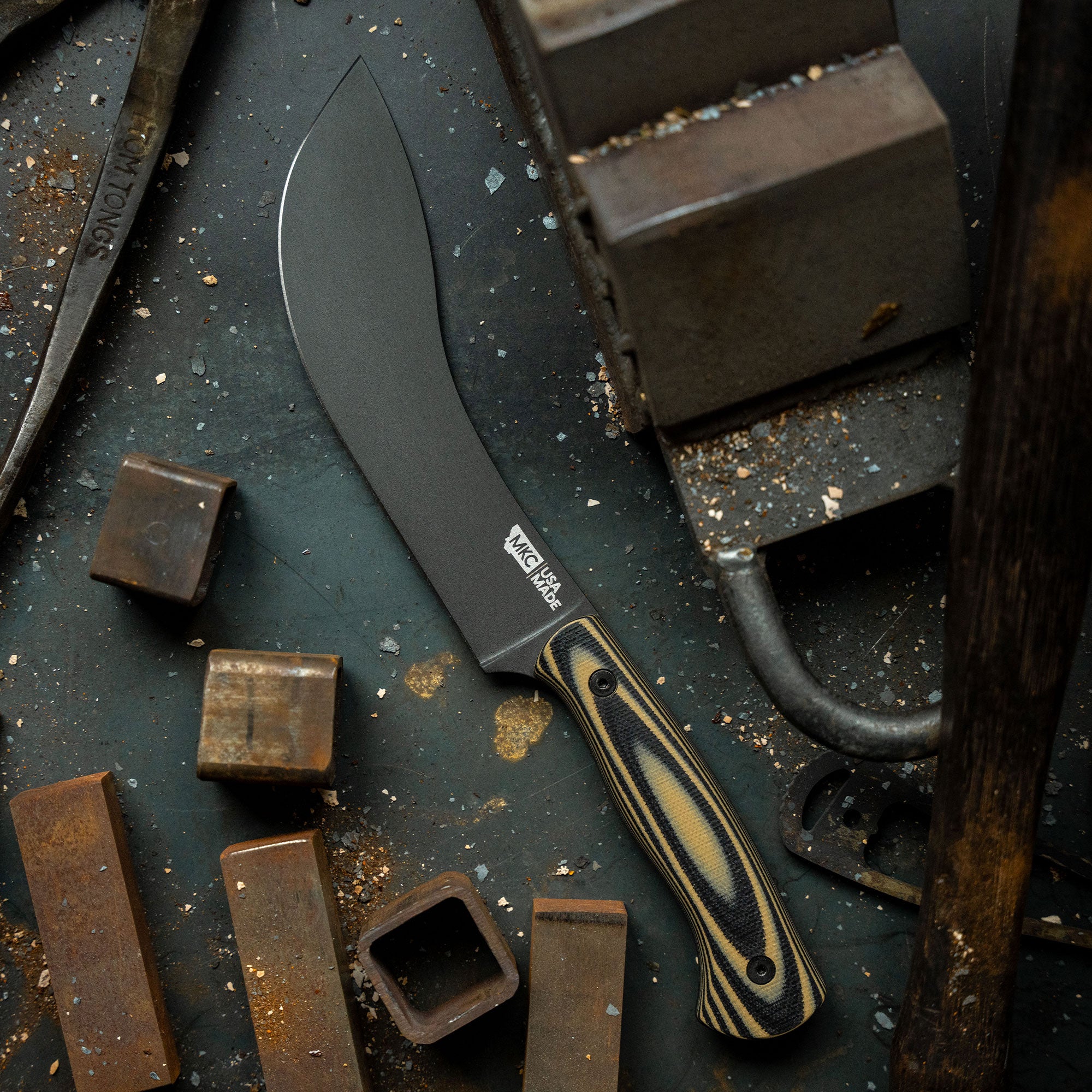 Alt text: Close-up of Beartooth Pro Skinning Blade with tan and black handle, featuring a matte black blade, displayed on a rustic dark metallic surface surrounded by scattered metal pieces and dust.