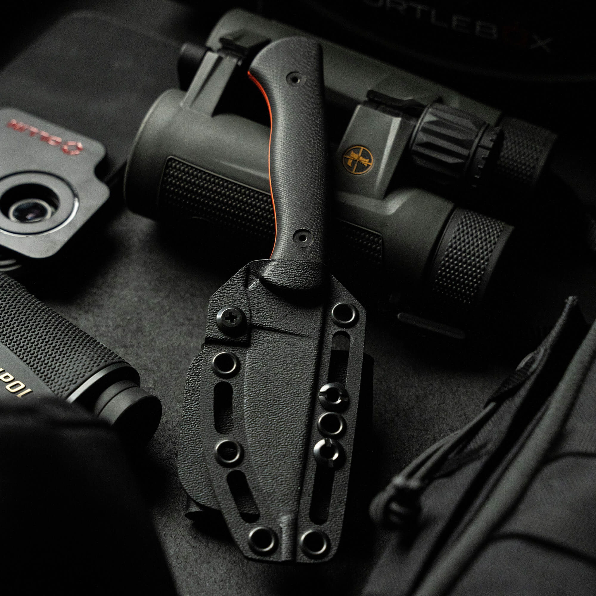 Alt text: "Close-up of the BLACKFOOT 2.0 knife in Blaze Orange Cerakote™ finish, displayed with its sheath amid various tactical gear including binoculars and other equipment, showcasing its sturdy design and bright accent along
