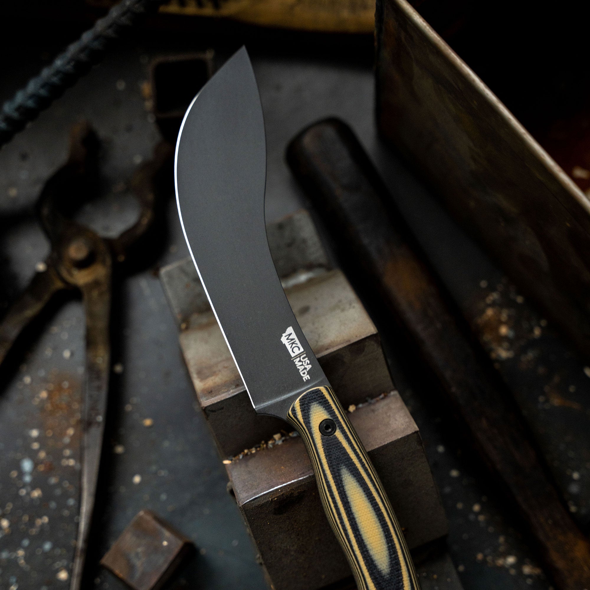 Alt text: Close-up of a Beartooth Pro Skinning Blade with a tan and black handle resting on a workshop bench, featuring a curved, matte black blade and the logo etched near the handle.