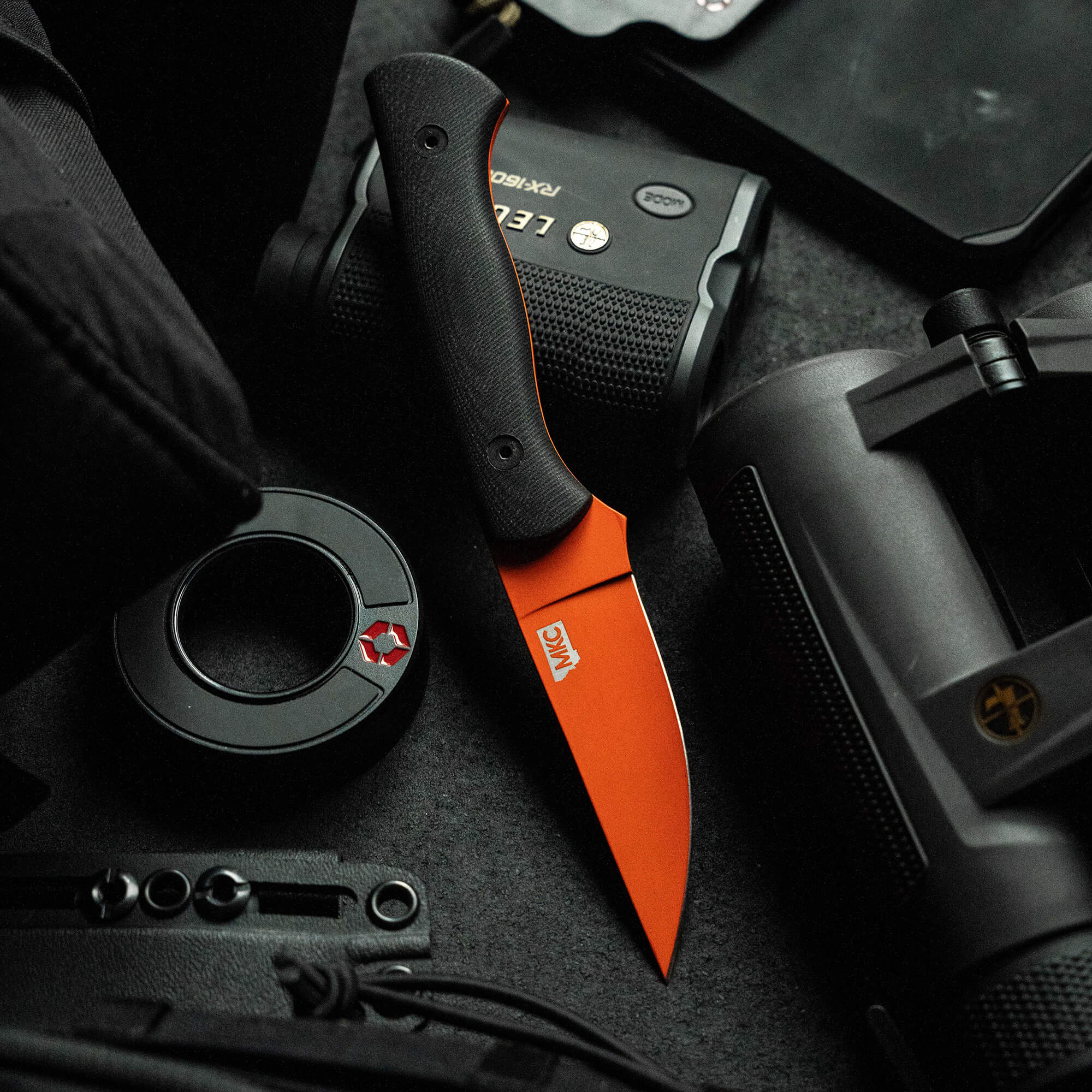 Alt text: "A BLACKFOOT 2.0 knife with a blaze orange Cerakote™ blade lies amidst tactical gear, including a camera, a rangefinder, and various black electronic devices, showcasing its sleek design and vibrant color contrast