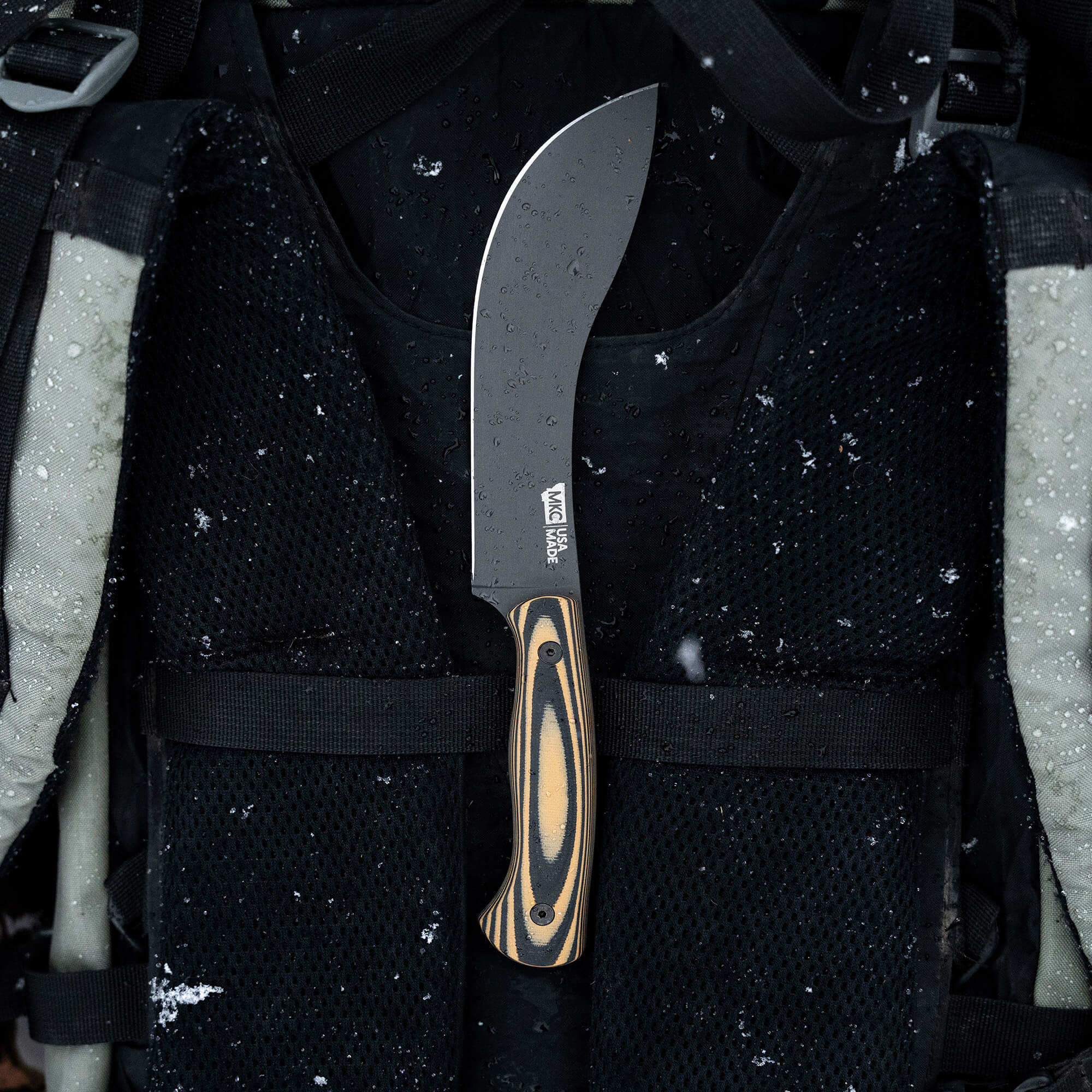 Alt text: "Beartooth Pro Skinning Blade with a tan and black handle attached to a tactical vest, covered in snowflakes, emphasizing the knife’s rugged use in outdoor environments."