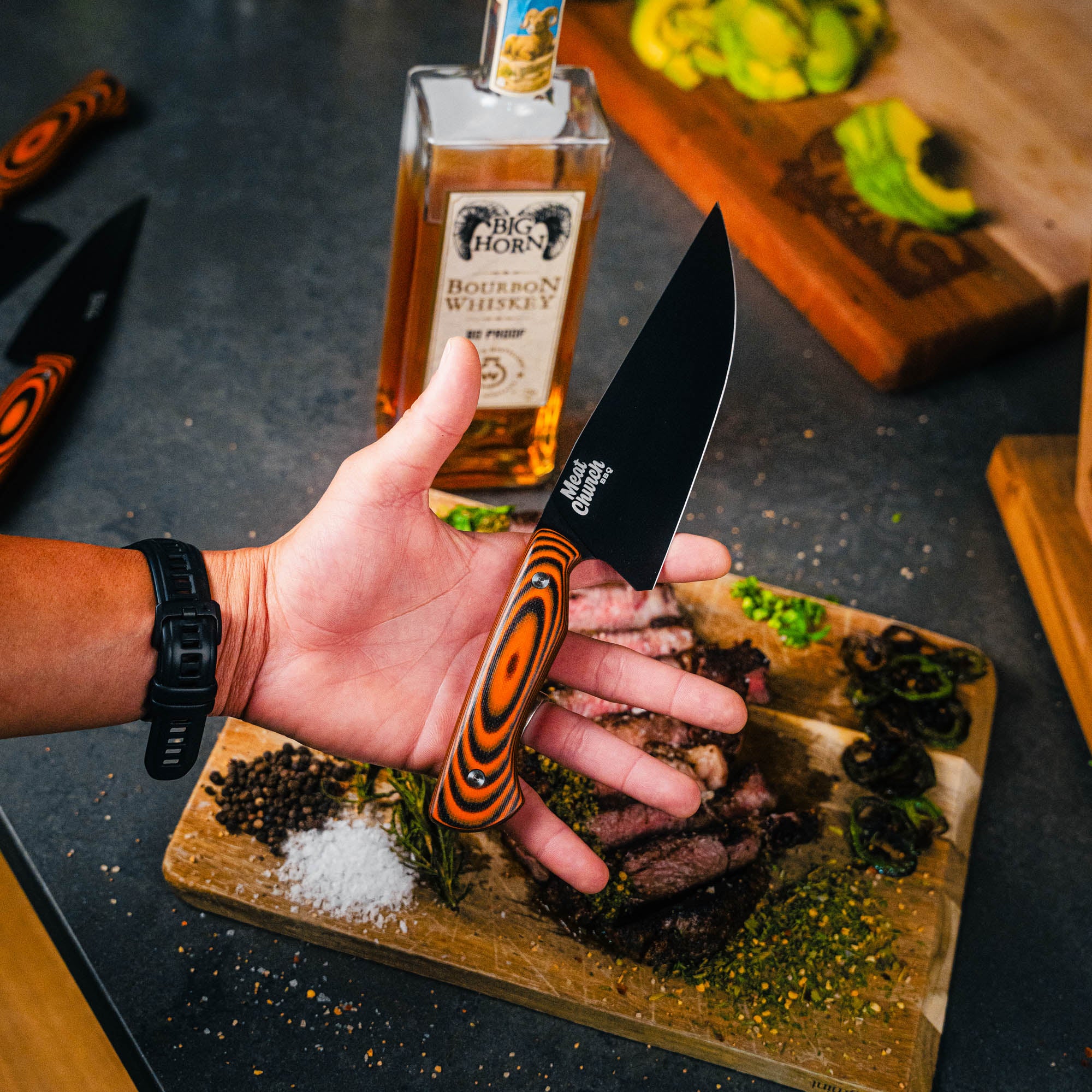 Meat Church chef knife with orange and black handle held over a cutting board with sliced meat and whiskey bottle.